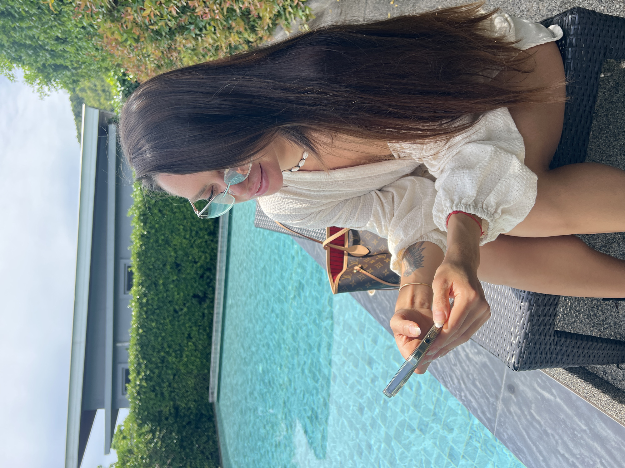 A woman with long, dark brown hair and light-colored sunglasses is sitting on a dark gray woven lounge chair next to a bright blue swimming pool. She is looking down at a clear phone case in her hands, which she is holding with both hands. Her left hand is resting on her right forearm, where a tattoo of a bird with spread wings is visible. She is wearing a white, textured, loose-fitting top with puff sleeves and a shell necklace. A Louis Vuitton Speedy bag with red interior lining is placed behind her on the lounge chair. The background features a neatly trimmed green hedge, a modern grey building with a sloped roof, and lush greenery. The scene appears to be outdoors in a tropical or subtropical climate, consistent with a location in Ban Bang Rak, Thailand.