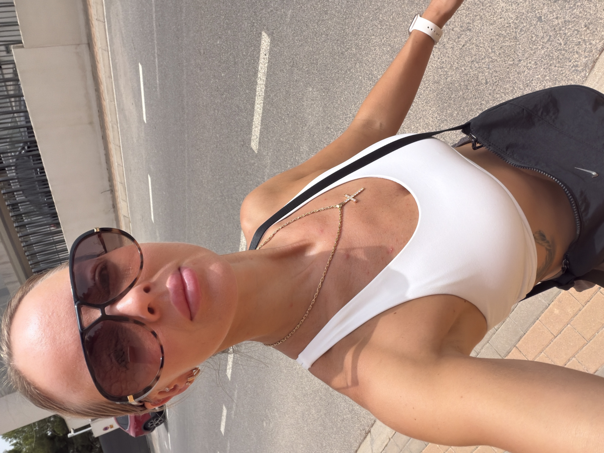 A woman stands on a paved sidewalk next to a street in Finestrat, Spain. She is wearing a white crop top, sunglasses, a gold chain with a cross pendant, and a black fanny pack. The street has white lane markings and a red car is visible in the background. The weather appears to be sunny.