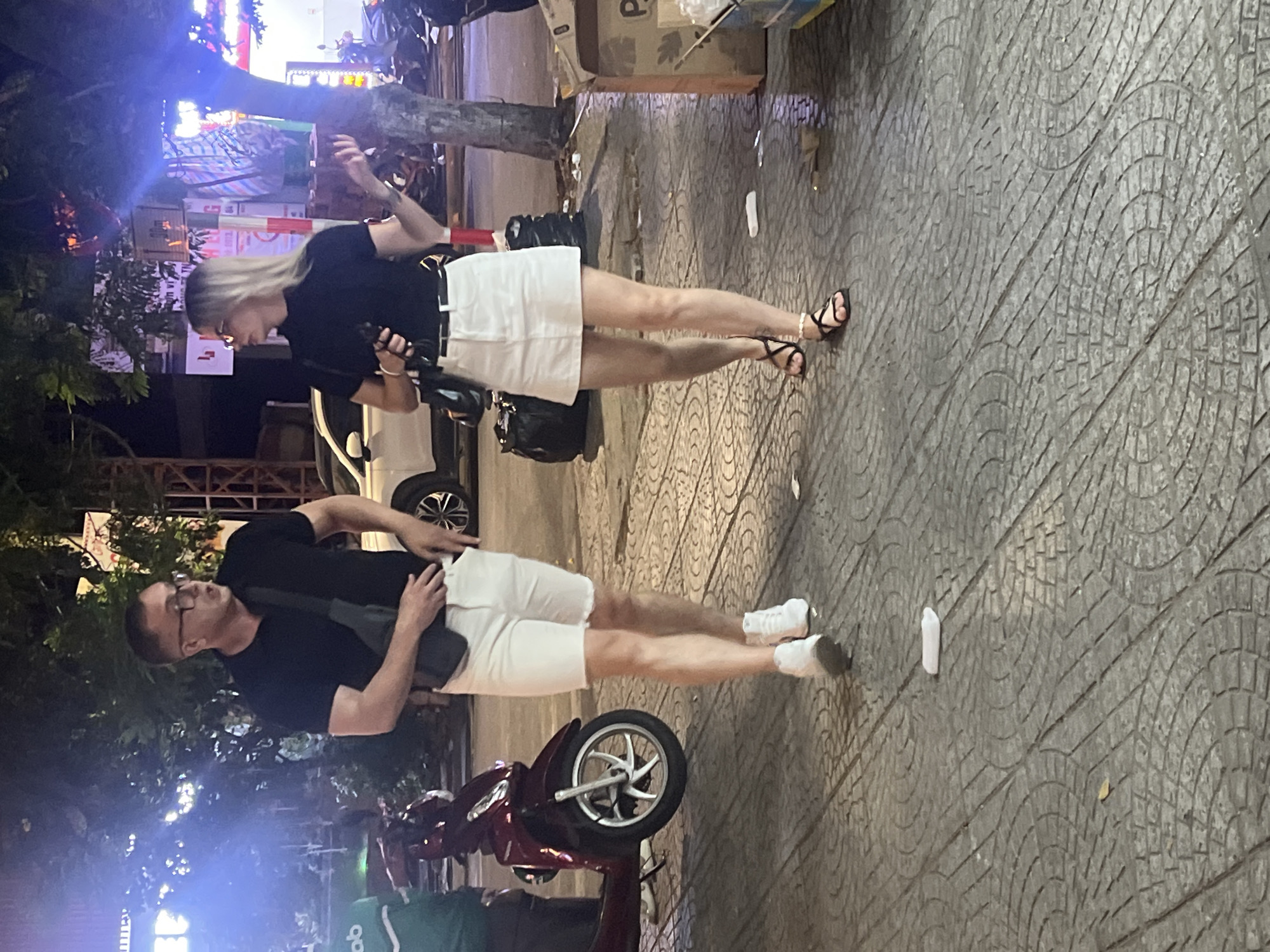 In Đà Nẵng, Vietnam, at night, two individuals are walking on a paved sidewalk. The person on the left, a man wearing glasses, a black t-shirt, and white shorts, carries a dark crossbody bag. The person on the right, a woman with blonde hair, wears a black t-shirt, a white skirt, and black sandals. Behind them, a red scooter is parked, and a white car is visible. The environment includes trees, streetlights, and various buildings in the background. A cardboard box and some litter are present on the sidewalk.
