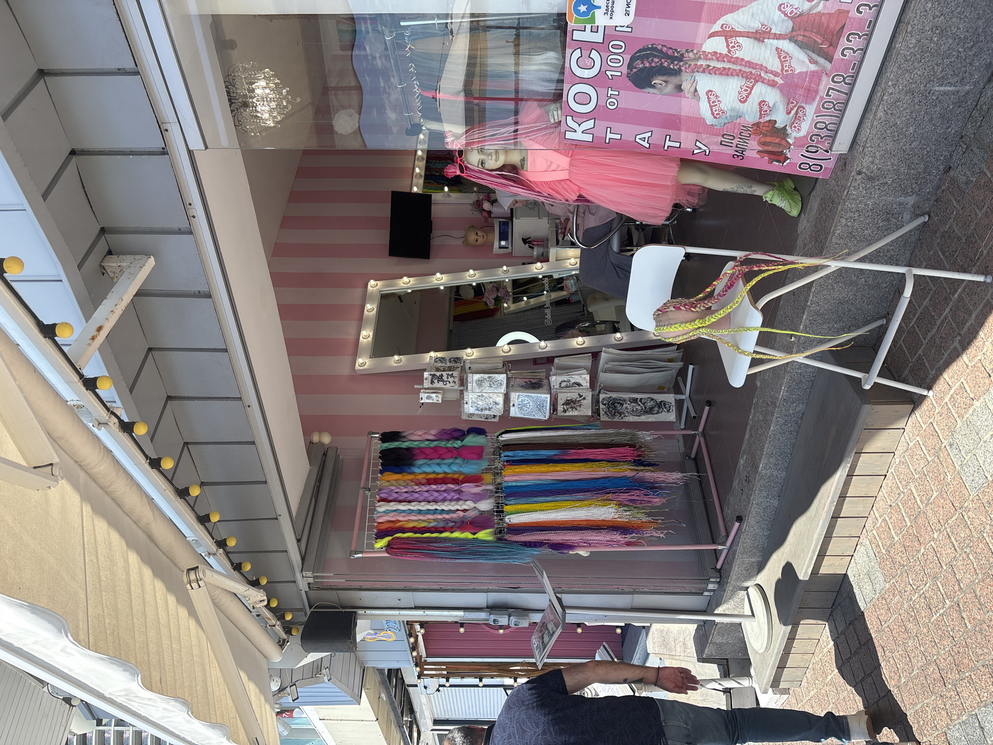 This media file captures the storefront and interior of a vibrant hair braiding salon in Sochi, Russia, during daytime.
**Subjects:** The image features a partial view of a man in a dark t-shirt and jeans walking on a brick sidewalk. Inside the shop window, a mannequin with long pink braids, dressed in a pink tutu-style dress and bright green sneakers, is prominently displayed. The shop's interior includes a large rectangular mirror framed with numerous light bulbs, a small black television screen, and a head mannequin. Two racks hold an extensive collection of colorful synthetic hair extensions or kanekalon braids, arranged in various hues including pinks, blues, greens, yellows, purples, and oranges. Above these racks are displays of smaller items, possibly temporary tattoos or hair accessories. A white folding chair inside the window holds several braided hair samples.
**Activity or event taking place:** The scene depicts a beauty salon specializing in hair braiding and possibly hair extensions, showcasing its services and products through its window display.
**Notable details:** The time of day is bright daylight, indicated by strong sunlight creating shadows on the brick sidewalk and a nearby stone bench. The shop's interior decor is characterized by a striking pink and white striped wall, a glamorous chandelier reflected in the window, and abundant lighting around the mirror, suggesting a lively and fashionable aesthetic. An awning with yellow string lights extends over the sidewalk.
**Visible text:**
On the large pink poster, Russian text includes "КОСЕ" (likely part of "КОСЫ" meaning "braids"), "от 100 к" (possibly "from 100 rubles"), "Т А Т" (likely "ТАТУ" meaning "tattoos"), and "ПО ЗАПИСИ" ("by appointment"). A phone number "8(238)878-33-3" is also visible. Small repeated text "Sichs" and "Sichsa" appears on the poster. In the mirror's reflection, an Instagram handle "@doll_house_sochi" is legible. Other partially visible text includes "Здесь хорошо" ("here is good"), "MODOX", and "2ГИС" (a Russian mapping service).