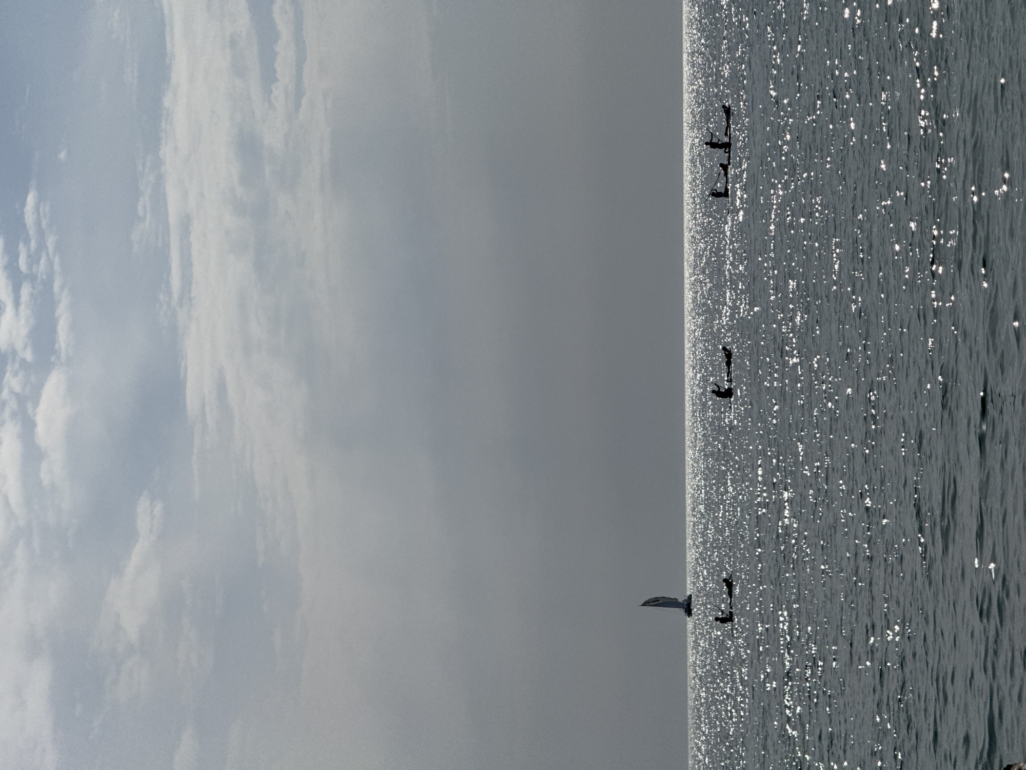 The image depicts a serene seascape with a hazy sky filled with scattered clouds. The sun, positioned high and slightly to the left, casts a brilliant, shimmering reflection on the calm water, creating a glittering effect across the surface. The horizon line is sharply defined, separating the muted blue of the sky from the dark, textured expanse of the sea.
In the mid-ground, a lone sailboat with its sail unfurled navigates the waters. To the right of the sailboat, several individuals are visible on stand-up paddleboards. One paddleboard has two figures, while others appear to have single occupants. They are silhouetted against the bright reflection, their forms distinct against the sparkling water. The overall atmosphere is peaceful and expansive, suggesting a tranquil day at sea. The image is taken from a low vantage point, looking out over the water towards the horizon.
While the image does not explicitly identify its location within Barcelona, Spain, the seascape and the style of recreational watercraft suggest a coastal setting typical of the city's extensive shoreline.