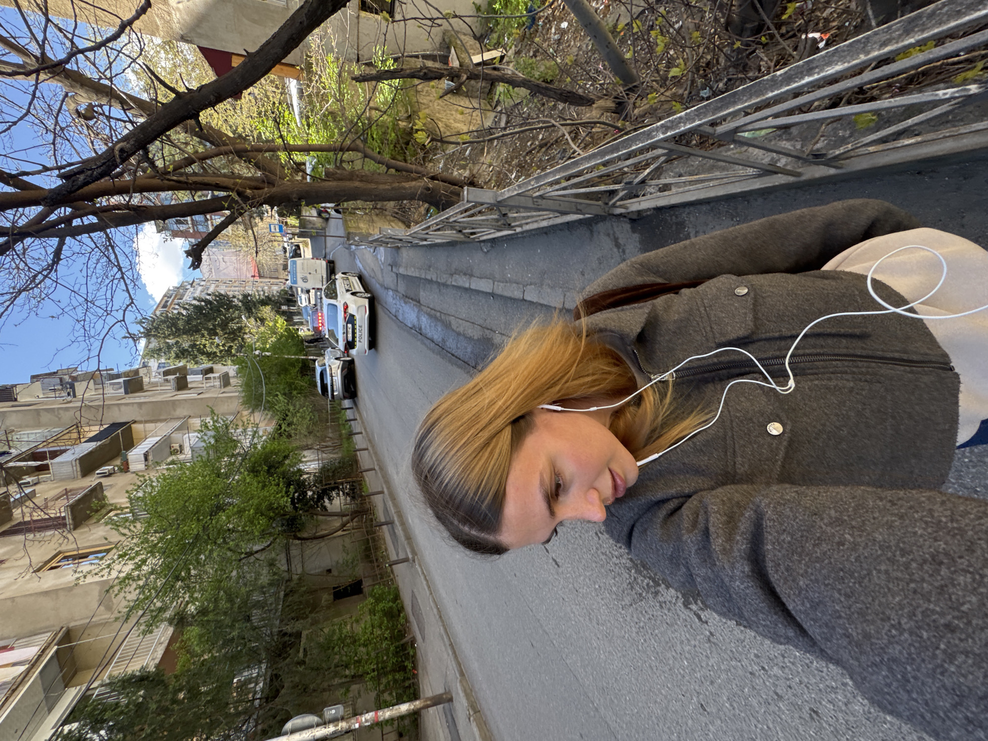 A woman is walking on the sidewalk of a street in Tbilisi, Georgia. She is wearing a gray coat and white headphones. Cars are parked along the side of the street. Trees and buildings are visible in the background. The street is paved and has a metal railing along the sidewalk.