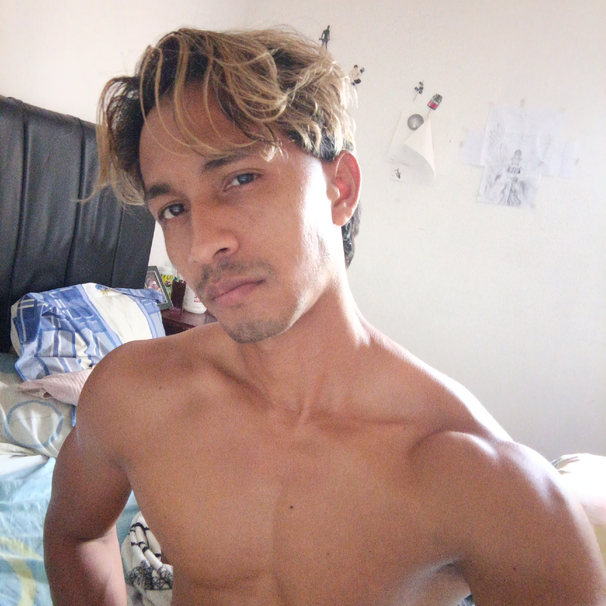 The image is a close-up, chest-up selfie of a shirtless man with light-colored, tousled hair. He is in what appears to be a bedroom, indicated by a bed with blue and white checkered bedding and a dark headboard. There are some papers and small figures attached to the wall behind him. The lighting suggests natural light coming from the right side of the frame. The man has a slight stubble and is looking directly at the camera. His left shoulder and upper chest are visible, showing some muscle definition. The overall impression is of a casual, private setting. The reference to "Piloncito, Venezuela" is not supported by any visual information within the image itself.