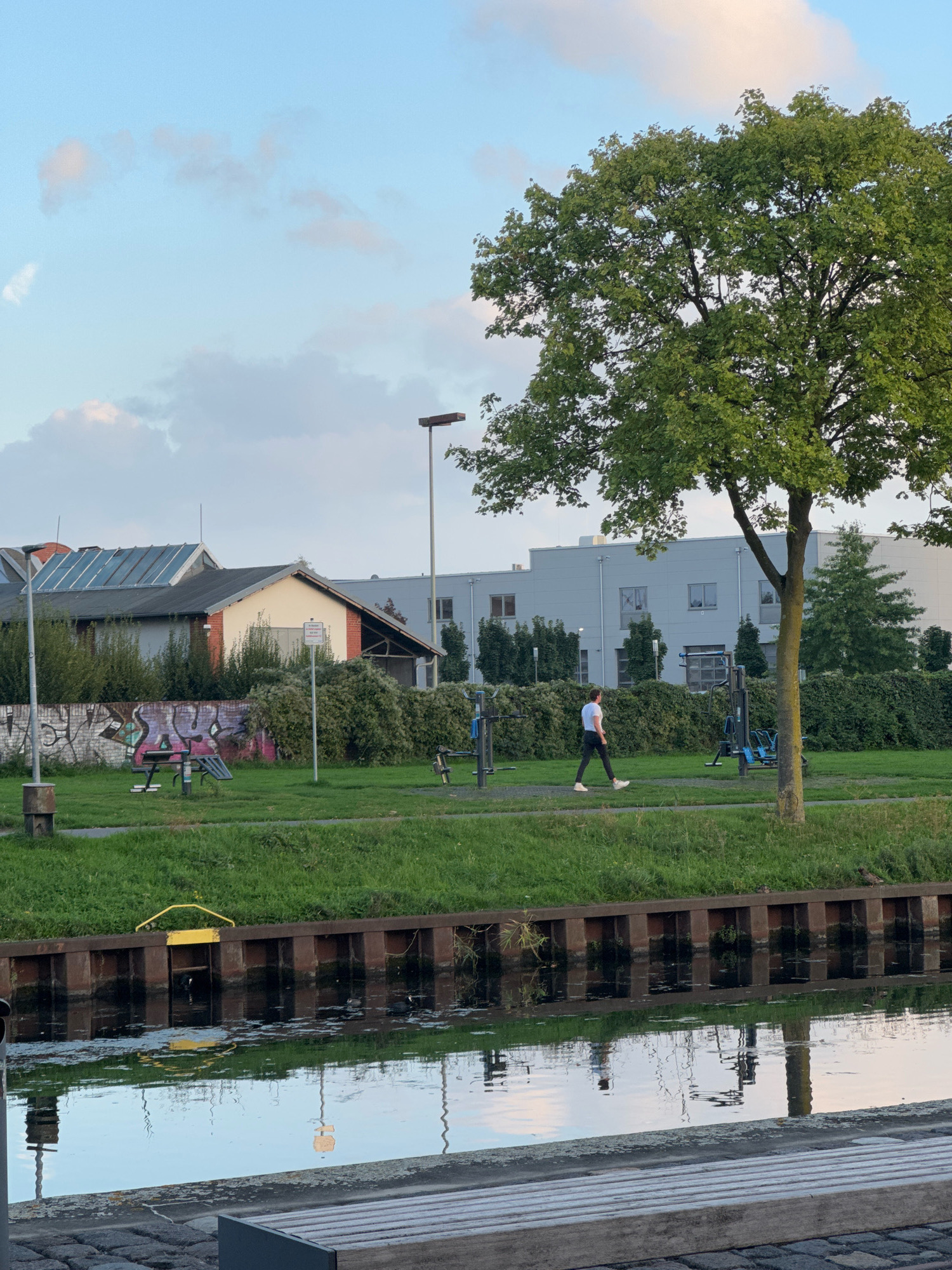 The image depicts an outdoor scene next to a canal, possibly in the city of Cleves, Germany, given the context. In the foreground, a wooden bench is visible on a cobblestone path, overlooking the dark, reflective water of the canal. The canal is bordered by a sturdy wooden and metal embankment.
Further back, a grassy area features several pieces of outdoor exercise equipment, including what appears to be a leg press machine and a pull-up bar. A person, wearing a white t-shirt and dark pants, is walking along a paved path in this exercise area. Behind the exercise equipment, a low brick wall with graffiti runs along the edge of the grass, backed by a dense green hedge.
Beyond the greenery, a building with a pitched roof and solar panels is situated, with a larger, modern, light-colored industrial or commercial building visible in the background. A tall lamppost stands near the building with solar panels. A large tree with lush green foliage is prominent on the right side of the image, casting a shadow. The sky is a pale blue with scattered clouds. The overall atmosphere suggests a peaceful, late afternoon or early evening setting in a semi-urban environment.