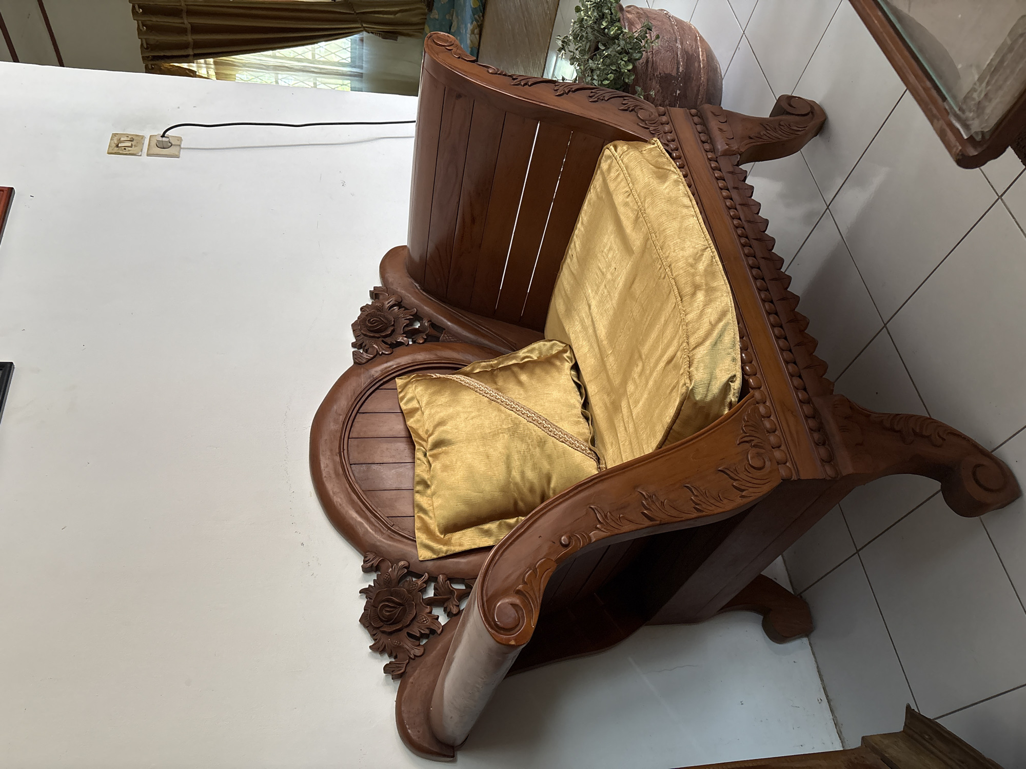 The image shows a carved wooden chair with gold cushions in what appears to be a living room in Uleepulo, Indonesia. The chair has ornate carvings of roses on the armrests and detailed decorative patterns along its base. A gold throw pillow rests on the seat cushion. To the right of the chair, a large potted plant with green foliage is visible, and further in the background, a window with gold curtains can be seen. A wall outlet with a black electrical cord plugged into it is mounted on the white wall behind the chair. The floor is tiled with large, light-colored square tiles. The overall scene suggests a domestic setting in a tropical climate.