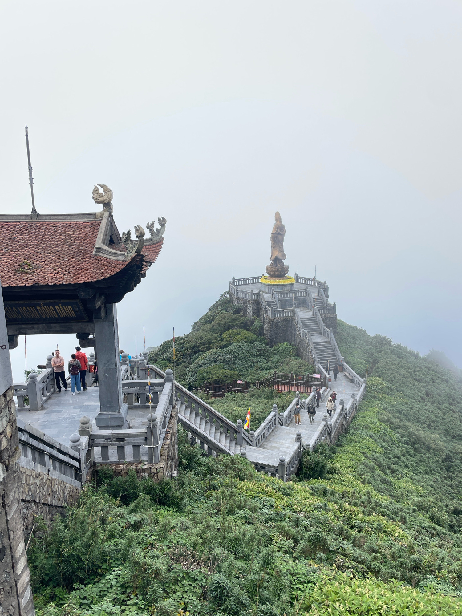 The image depicts a mountainous landscape in Vietnam, possibly Sa Pa or Sapa, on a foggy day. In the foreground, a traditional Vietnamese-style temple structure with a red tiled roof and ornate carvings is visible. Several people are gathered on a stone pathway in front of the temple. A sign with Vietnamese text is mounted on the structure.
Further into the scene, a wide stone staircase ascends a lush green mountainside, leading to a large plaza. Atop the plaza stands a towering golden statue of a female figure, likely a deity or a revered person. The statue is intricately detailed and appears to be made of bronze or a similar material. The plaza is surrounded by stone walls and decorative elements.
The mountainside is densely covered with green vegetation, with trees and shrubs clinging to the steep slopes. The mist or fog creates a hazy atmosphere, obscuring the distant landscape and adding a sense of ethereal beauty to the scene. Several people can be seen walking up and down the staircase, suggesting that this is a popular tourist destination or a place of pilgrimage. The overall impression is one of grandeur and serenity, with the impressive architecture and natural beauty blending together.