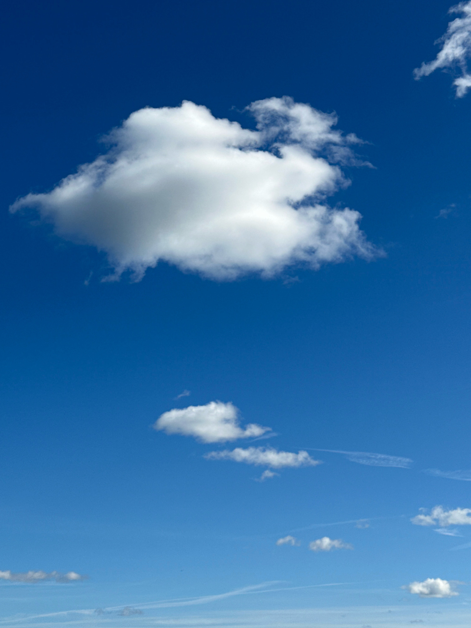 This image displays a bright blue sky with scattered white clouds. The main subject is the sky itself, with a large, fluffy cumulus cloud dominating the upper left portion of the frame. Below it, two smaller cumulus clouds are visible, with wispy contrails stretching across the sky in the distance. The overall scene is serene and peaceful.
The time of day appears to be daytime, given the bright sunlight illuminating the clouds and the clear blue sky. The weather seems fair and pleasant, with only scattered clouds and no indication of rain or storms. There are no people or man-made objects visible in the frame, suggesting an outdoor, natural setting. The location is identified as Tallinn, Estonia, though there are no specific landmarks or architectural cues to confirm this within the image itself. The image evokes a sense of calm and vastness, common emotions associated with looking up at a clear sky. There is no visible text in the image.