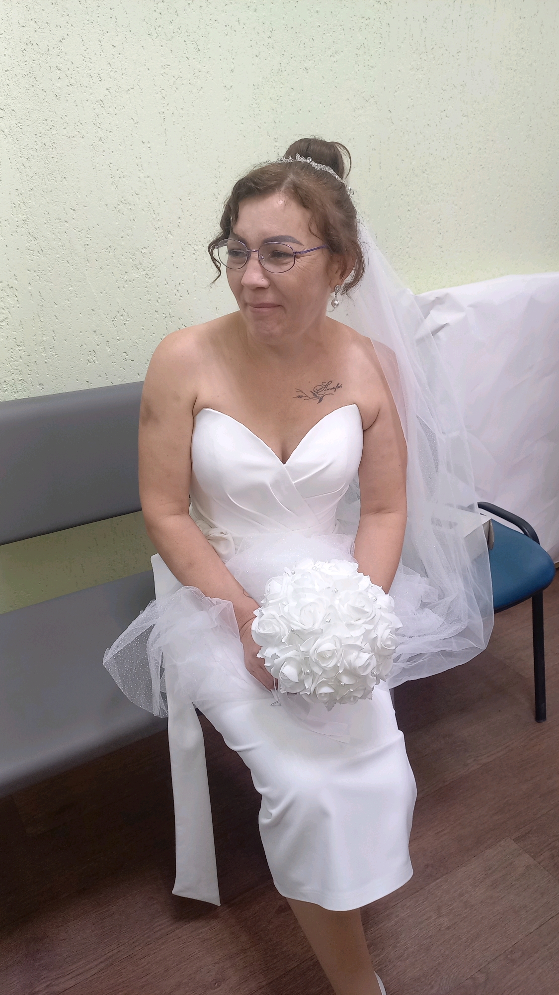A woman, appearing to be a bride, is seated on a gray bench against a light green textured wall. She is wearing a white strapless wedding dress and a veil. She has her hair pulled up with a tiara and is wearing glasses and pearl earrings. She is holding a bouquet of white roses. A tattoo is visible on her upper chest. The floor is dark wood. The overall scene appears to be indoors.