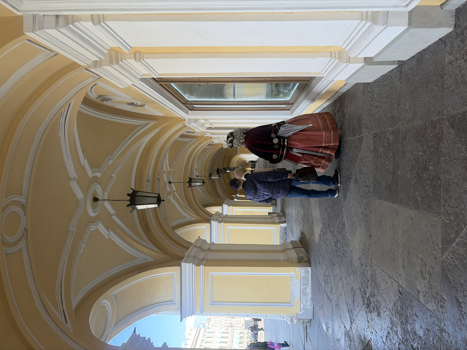 The image captures an ornate arched corridor or colonnade, likely part of a grand historical building in Saint Petersburg, Russia. The ceiling is vaulted and features intricate white decorative molding against a pale yellow background, from which several large, dark metal lanterns are suspended. The walls are also painted a light yellow with white architectural trim, creating a bright and elegant atmosphere. The floor is paved with dark gray stone tiles.
In the mid-ground, two individuals are prominent. On the right, a person is dressed in elaborate traditional or historical attire, characterized by a full red skirt with intricate patterns, a dark velvet-like cloak or jacket, and a distinctive hat or headpiece. This figure appears to be facing the other person. Opposite them stands an individual in modern casual wear, including a dark blue padded jacket, blue jeans, and a baseball cap. They seem to be observing or interacting with the traditionally dressed person, possibly a performer, guide, or historical re-enactor.
The corridor extends into the background, showcasing more arches and structural details. Bright daylight illuminates the scene, suggesting it is daytime, and light reflects off a window situated in the upper part of the wall. The overall scene depicts a casual encounter or a moment of observation within a historically rich architectural setting. There is no visible text in the image.
