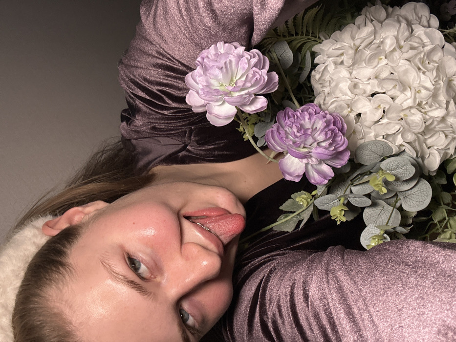 A person lies on their side, sticking out their tongue. They wear a plush headband and a shiny, dark purple, textured top. Two light purple flowers and a white hydrangea are positioned above them, intertwined with various green leaves and foliage. The background is a plain grey.