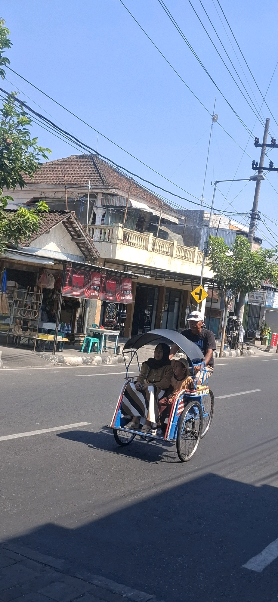In Blitar, Indonesia, a rickshaw carrying three people travels along a paved road under a clear blue sky. The rickshaw is pulled by a man wearing a dark t-shirt and a white cap, who is seated at the rear. In front of him, a woman wearing a hijab and a brown outfit is seated, holding a young child who is also wearing a brown outfit and a hat. The rickshaw has a black canopy and is blue with red and white accents. The road is bordered by a sidewalk and a building with a tiled roof, shops with banners, and trees. Electrical wires are visible overhead, strung between poles. A yellow traffic sign with a t-junction symbol is mounted on a pole near the sidewalk. The scene appears to be a daytime shot, with shadows cast by the rickshaw and overhead wires.