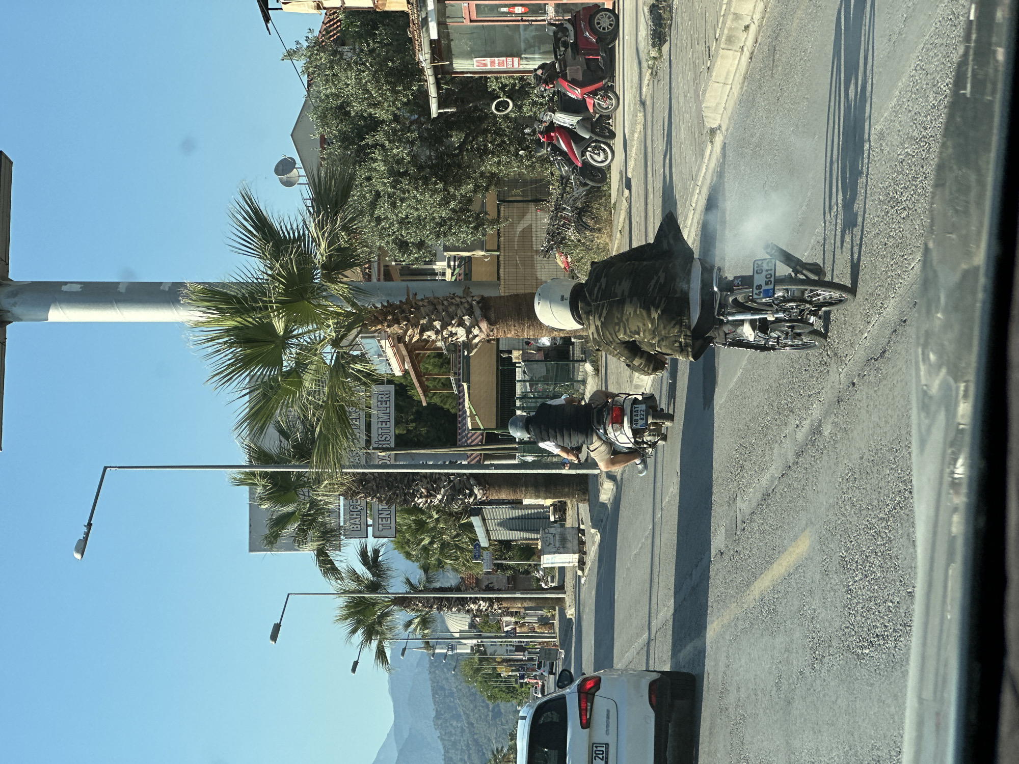 The image depicts a street scene in Fethiye, Turkey. A white car with the license plate "34 EAU 201" is stopped on the road. A motorcyclist with a visible license plate "48 AAT 822" is driving on the right side of the road and is passing by palm trees, street lights, and buildings. There is another person wearing a military-style jacket standing near an overturned motorcycle. Smoke is coming out from beneath the overturned motorcycle. Another row of motorcycles can be seen in the distance behind the first overturned motorcycle. Mountains are visible in the background.