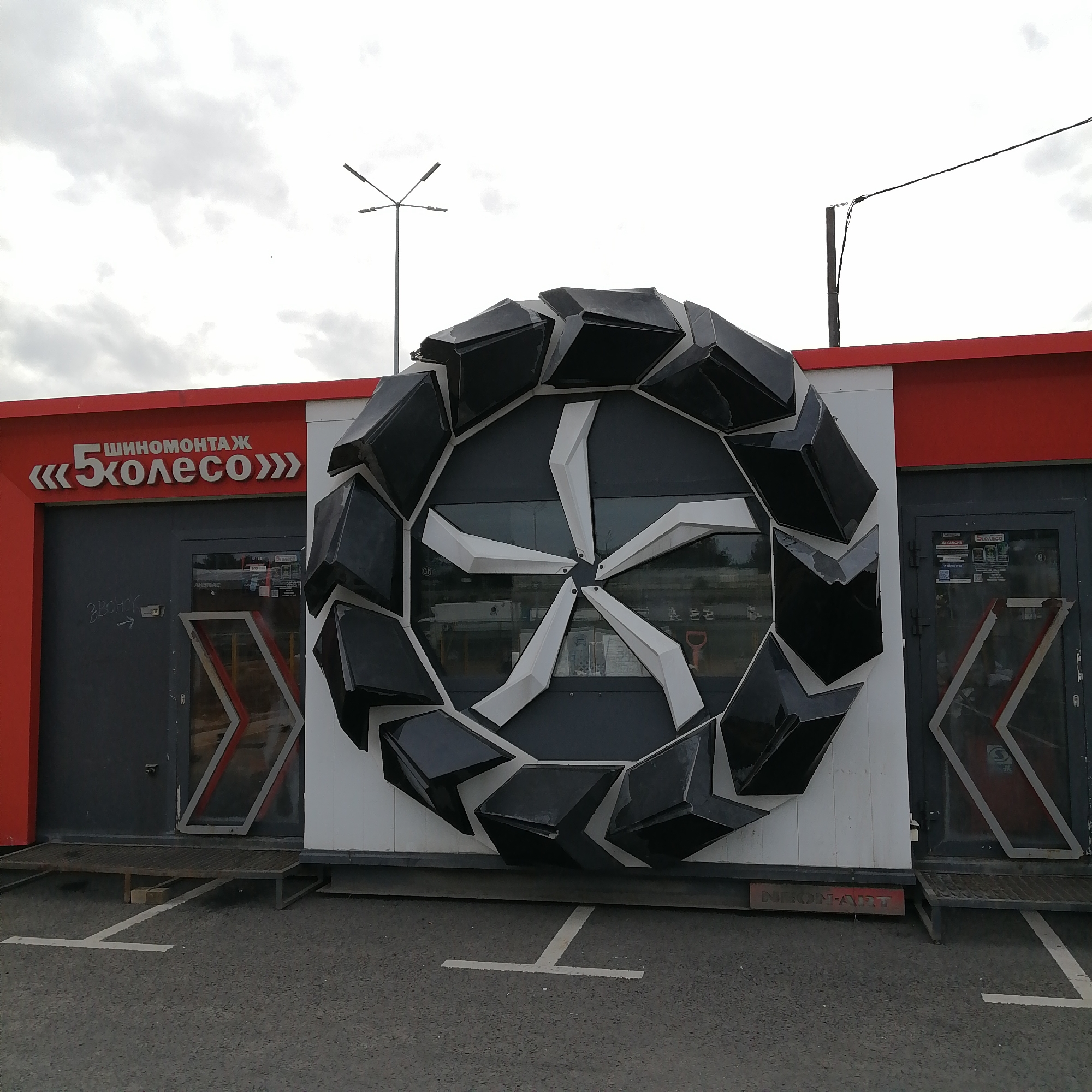 The image displays the exterior of a commercial building, likely a tire service shop, on an overcast day. The structure is a single-story, modular unit featuring a red top band and dark gray lower sections, separated by a white central panel.
A large, three-dimensional sculpture of an abstract wheel or tire is mounted prominently on the white central section of the building. This artwork is composed of black, angular, block-like segments forming the outer rim, and white, multi-faceted spokes radiating from the center. The spokes appear to be made of similar angular forms.
On the red section of the building to the left, white text reads "ШИНОМОНТАЖ" (Tire Fitting) and "«5 КОЛЕСО»" (5th Wheel), identifying the business. Below this signage is a dark gray door with a glass panel, displaying some faint graffiti or text, potentially "ВЫСУШ" (Dry Out) with an arrow. Another similar dark gray door with glass is visible to the right of the wheel sculpture, adorned with white and red chevron patterns. Near the base of the building, on the right, a small red sign bears the text "NEON-ART."
The foreground consists of an asphalt pavement with white 'T'-shaped markings, indicating parking spaces. A metal gridded ramp or step provides access to the building's doors. In the background, under a cloudy, light gray sky, a multi-armed street light pole is visible on the left, and a black electrical wire extends from a pole on the right. No people or specific activities are depicted; the scene is stationary, focusing on the architecture and signage of the business.