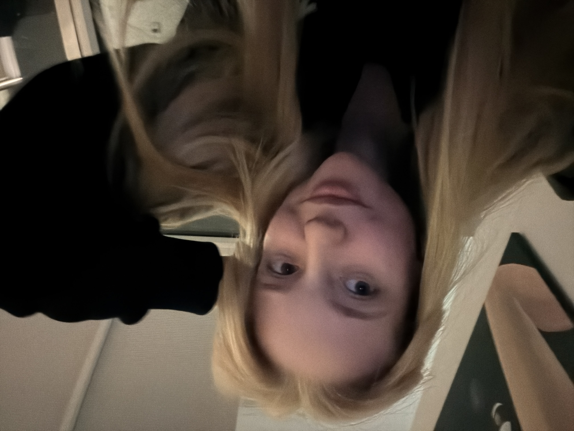 A portrait of a blonde-haired person is presented upside down. The individual's face is visible, with discernible facial features. The person is wearing a black garment, possibly a hoodie or jacket. The background contains indistinct elements, including what appears to be a wall and a framed object. The location and specific actions are unidentifiable based on the image alone.