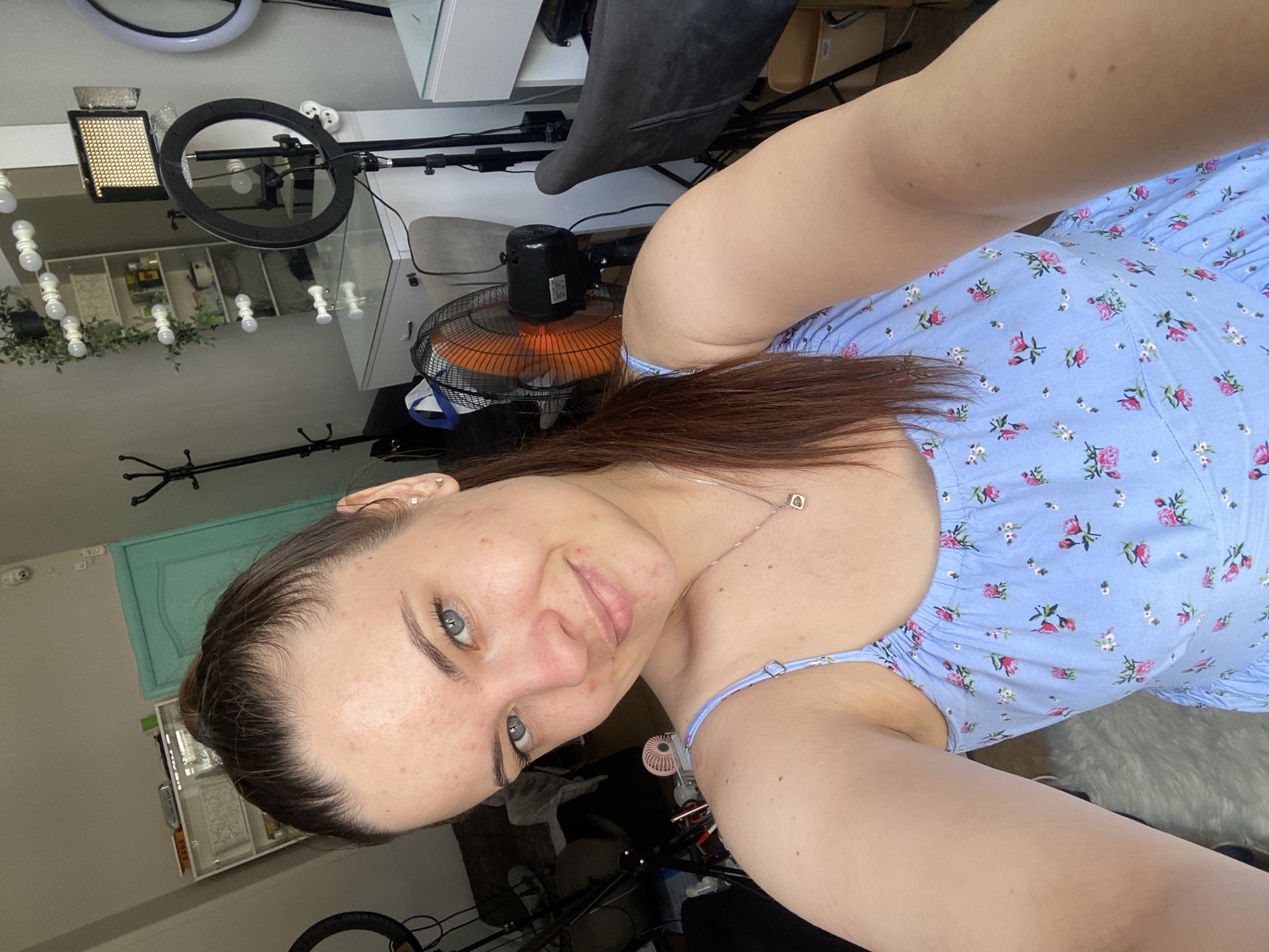 The image is a selfie taken by a young woman with brown hair pulled into a high ponytail. She has light blue eyes and is smiling slightly. She is wearing a light blue dress with a floral pattern of pink and white flowers. Her skin appears to have some blemishes. In the background, there is a room that appears to be set up for filming or content creation. There is a vanity mirror with lights, a ring light, a softbox light, a fan, and a coat rack. A teal colored door is visible in the background. The image does not contain any information that would allow for its localization to Poltava, Ukraine.