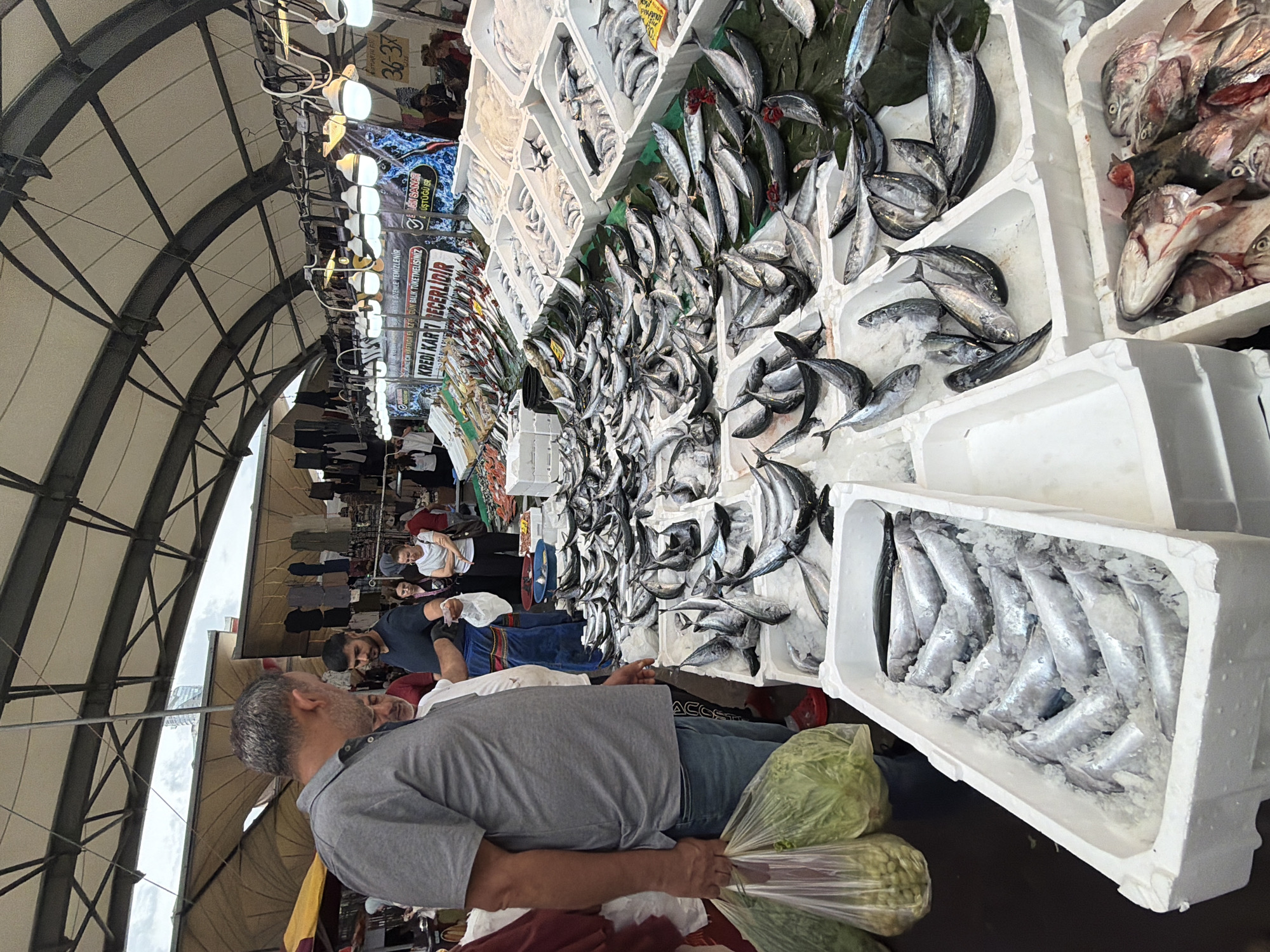 The image depicts a bustling fish market, likely in Kadıköy, Turkey, given the context of the prompt. In the foreground, a man in a grey polo shirt and blue jeans carries two large plastic bags filled with green produce. He is looking towards a stall displaying a wide variety of fresh fish laid out on ice in white styrofoam boxes. Several other people are visible browsing the fish stalls. In the background, the market is covered by a large, arched canopy structure with visible metal beams and lighting fixtures. Signs in Turkish are present, indicating prices and possibly information about the fish. The overall scene is one of active commerce, with abundant seafood for sale in an open-air market environment.