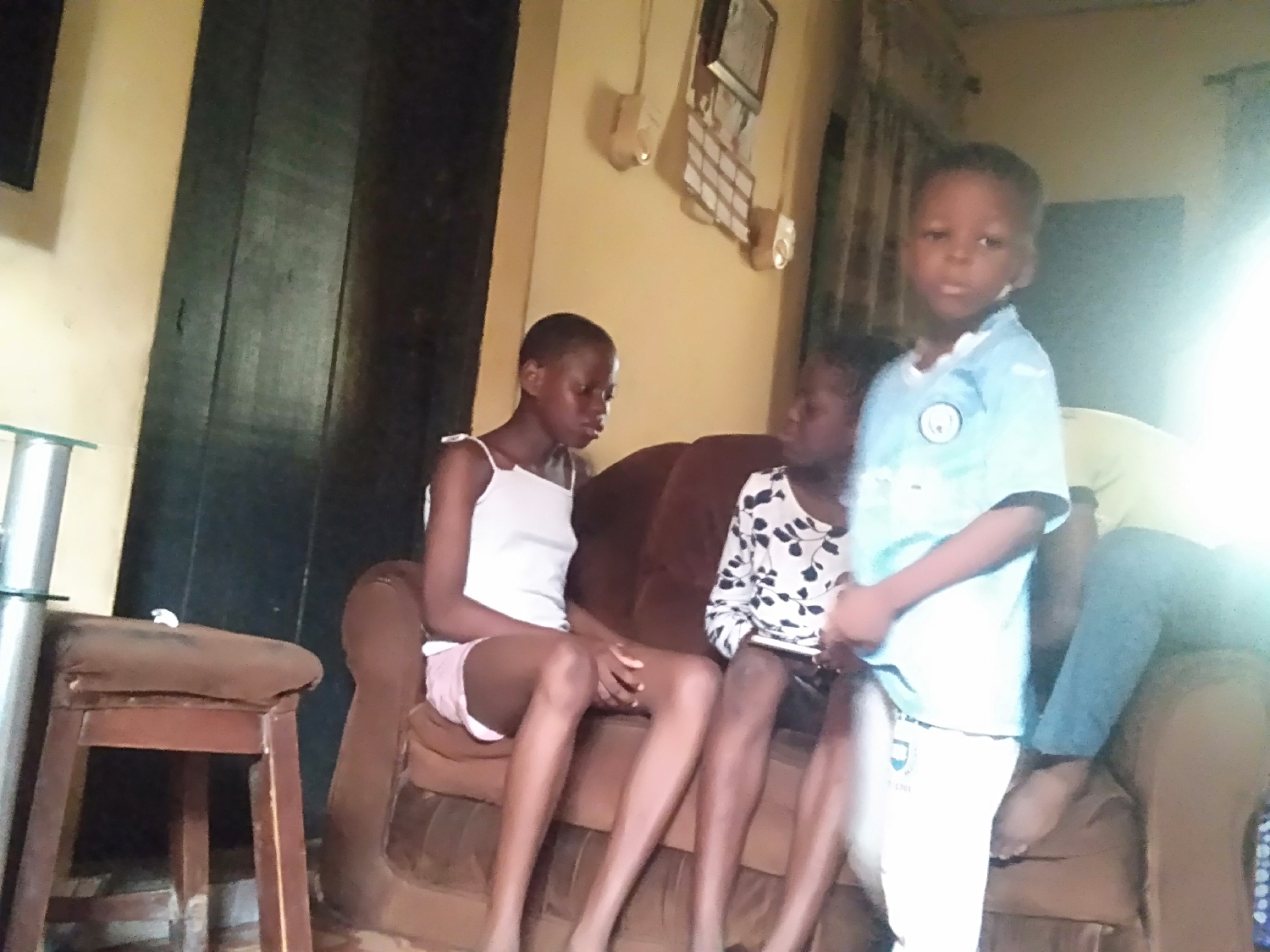 This image captures an indoor scene featuring multiple individuals, primarily children and young adults, gathered in what appears to be a living room or common area.
In the foreground, a young boy stands, facing the camera with a neutral expression. He wears a light blue t-shirt with a white circular logo on the left chest, resembling a football club emblem, and white pants that also bear a similar logo on the left thigh.
To the boy's left, a girl with short hair is seated on a brown sofa. She is dressed in a white spaghetti-strap top and light-colored shorts, looking downwards or to her side. Another person is partially visible sitting next to her on the same sofa. To the far right, another individual is seated, wearing a dark top with white patterns and blue jeans, their form partially obscured and overexposed by bright light.
The setting includes a brown upholstered sofa occupying the center of the frame. To the left, a small wooden stool or table with a metal base and a clear glass top is present. The background features a large, dark wooden door or partition, and the walls are painted a light beige or yellow. A framed item, possibly a picture or calendar, is visible on the upper left wall.
The scene is illuminated by strong, natural daylight entering from the right, causing glare and overexposure in that section of the image. The lighting suggests it is daytime. The activity appears to be a casual gathering, with individuals relaxing. No explicit text is clearly legible, though logos are present on the boy's clothing.