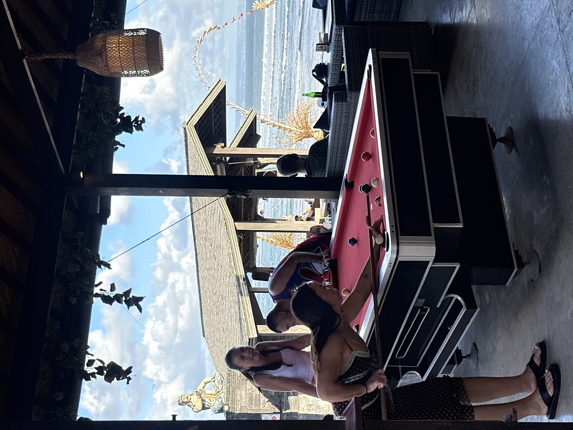 A group of people are gathered around a pool table at what appears to be an outdoor bar or lounge with a view of the ocean. The scene is set in North Kuta, Indonesia, suggested by the traditional Balinese decorations, including a spirit house or offering and ornate wooden structures. The time of day is likely daytime, with bright sunlight illuminating the sky and casting shadows. The weather appears to be pleasant, with a clear blue sky dotted with fluffy white clouds. The ocean in the background shows moderate waves, indicating a lively beach scene, possibly with surfers.
Several individuals are actively engaged with the pool table. One woman is reaching out with a pool cue, preparing to take a shot. Another woman stands beside her, looking on. A man is leaning over the pool table from the opposite side, also seemingly involved in the game. In the background, more people are visible, some seated at tables and others near the beach.
The setting is a tropical environment, with lush greenery and elements of traditional Indonesian architecture, such as thatched roofs and intricate carvings. A decorative woven lampshade hangs from an overhead beam, adding to the ambiance. The pool table itself has a red felt surface and is set up on a concrete floor. The overall mood seems relaxed and social, with people enjoying leisure time by the sea. There is no visible text in the image.