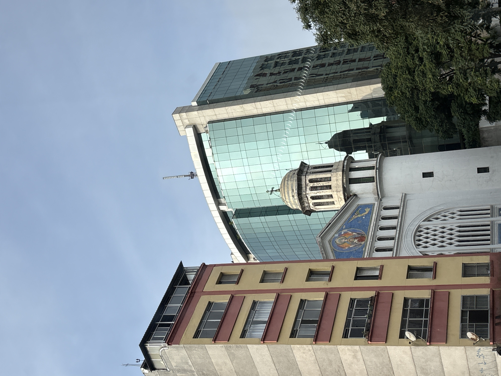 This is a low-angle, full shot of an urban street scene in São Paulo, Brazil, under a clear blue sky. In the foreground, on the left, is a tall, narrow apartment building with a beige and yellow facade, accented with maroon trim. The building has multiple floors with rectangular windows, some covered by curtains and others showing open blinds or empty interiors. Two satellite dishes are mounted on the side of this building. To the right of the apartment building is a white church with a large, arched entrance featuring intricate lattice-like window panes. The church has a dome topped with a cross. Above and behind the church is a modern, multi-story glass skyscraper with a light blue-green tint. The skyscraper's reflective surface mirrors the church and other buildings. A large green tree with dense foliage partially obscures the right side of the skyscraper. The scene appears to be from a city street, with elements of older architecture (the apartment building and church) juxtaposed with modern urban development (the glass skyscraper).