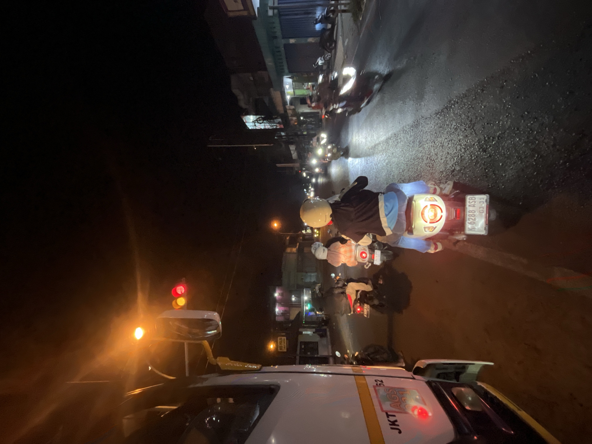 The image depicts a nighttime street scene in Welahan, Indonesia, likely near a traffic light, given the presence of a red and yellow traffic light visible above a white truck in the foreground. The truck, with markings "JKT AGS 021 52", is partially visible on the left side of the frame. The road is wet, reflecting the lights of various vehicles. A group of motorcycles, each with a rider and some with passengers, are on the road, suggesting a traffic jam or a queue at the intersection. One motorcycle in the foreground has a license plate reading "K 6288 ASB 03-21". Buildings with illuminated windows and signs line the right side of the street, indicating commercial establishments. The overall atmosphere is one of a busy, urban night scene.