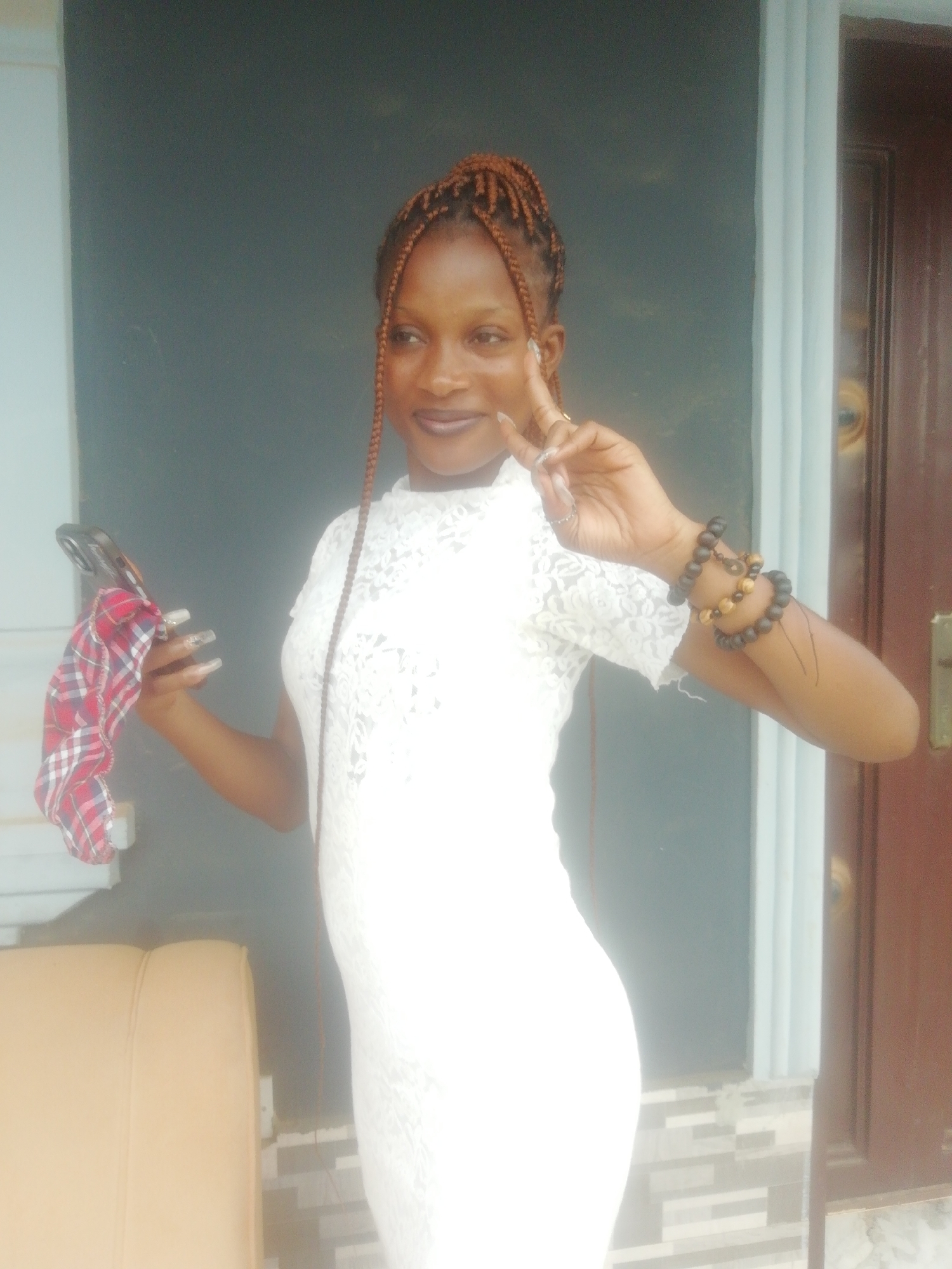 A young woman is captured in a full-body shot, posing for the camera with a subtle smile and making a peace sign with her right hand. She has dark skin and reddish-brown braided hair, styled upwards with some strands framing her face. She is wearing a white, short-sleeved lace dress with a high neckline.
Her left hand holds a smartphone, partially covered by a red and white plaid fabric, possibly a handkerchief or scarf. Her nails are long and manicured, and she wears dark lipstick. On her right wrist, she wears multiple beaded bracelets in dark and light brown tones.
The background features a dark gray wall on the left, and a lighter gray/blue vertical trim or frame on the right, adjacent to a dark brown door. Below the trim, a section of patterned tile flooring is visible. In the bottom left corner, part of a light brown/tan armrest, possibly from a sofa or chair, is visible. The lighting suggests it is daytime. The overall impression is calm and poised. There is no visible text in the image. The location context provided is Ikorodu, Nigeria.