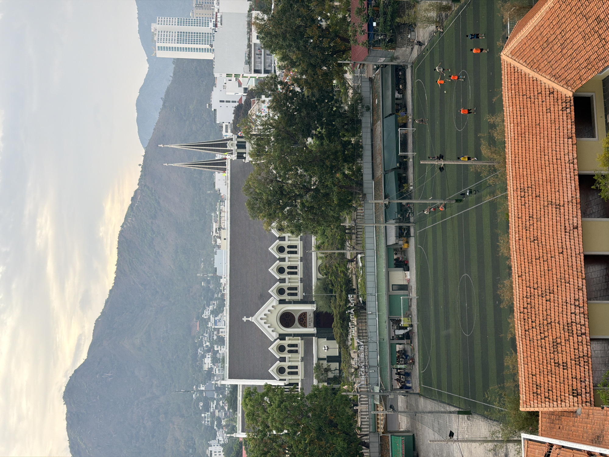 This aerial shot shows a cityscape in Bac Nha Trang Ward, Vietnam, with a mixture of natural and urban elements. A mountain range with partial cloud cover dominates the left side of the image. A church with a gray roof and twin spires is prominently featured near the center of the frame. Adjacent to the church is a football field where several individuals are engaged in a game. A building with a red tile roof is visible on the right side of the image. The background contains high-rise buildings and other urban structures. Trees are scattered throughout the scene.