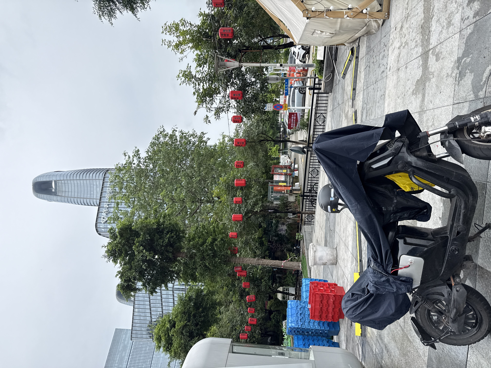 The image depicts an outdoor scene in the city of Haizhu District, China. In the foreground, a black electric scooter is covered with a black tarp. A black helmet rests on the back of the scooter. To the left of the scooter are stacks of blue and red plastic crates. Behind the scooter, red lanterns are strung across trees, and a tall, modern skyscraper with a distinctive curved top dominates the background. Several other glass-fronted buildings are visible, along with more trees and a street with parked cars. A white tent structure with wooden supports is present on the right side of the frame. The ground is paved with grey tiles. The sky is overcast.