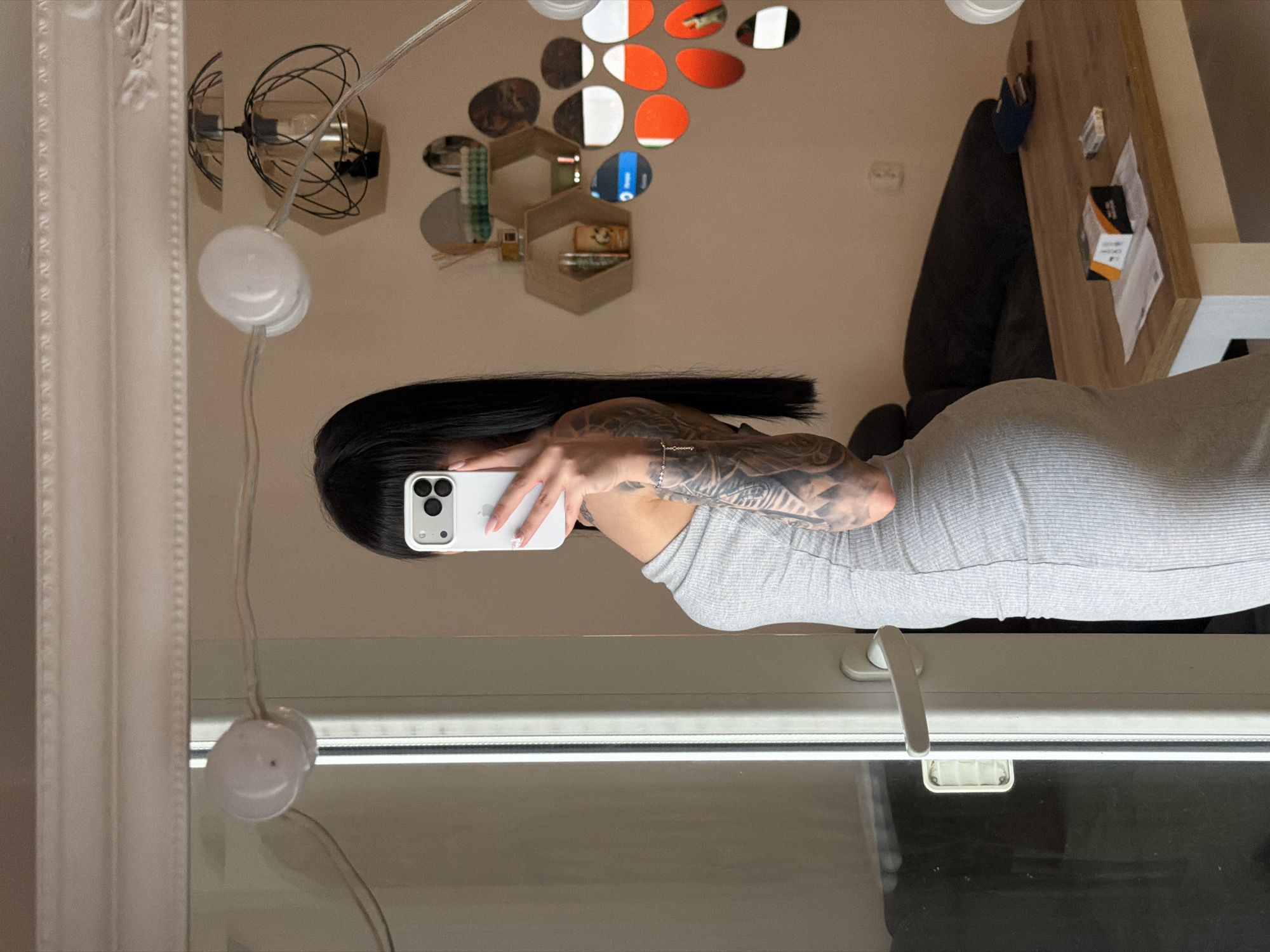A woman with black hair and tattoos on her arm is taking a mirror selfie in an apartment. She is wearing a gray, form-fitting dress. The mirror reflects a room with shelves on the wall decorated with circular mirrors and objects. A table with a box and other items is also visible. The scene is dimly lit. The location of this image is not Plovdiv, Bulgaria.