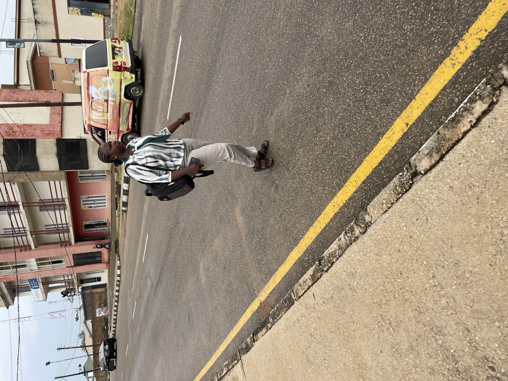 A woman is walking across a paved road in Jos, Nigeria, with a backpack slung over her right shoulder and her left hand holding a cellphone. She is wearing a striped short-sleeved shirt, light-colored pants, and sandals. The road has a double yellow line marking. In the background, there is a multi-story building with a pink facade, a parked yellow and orange van, and a black car on the left side of the road. The sky is overcast.