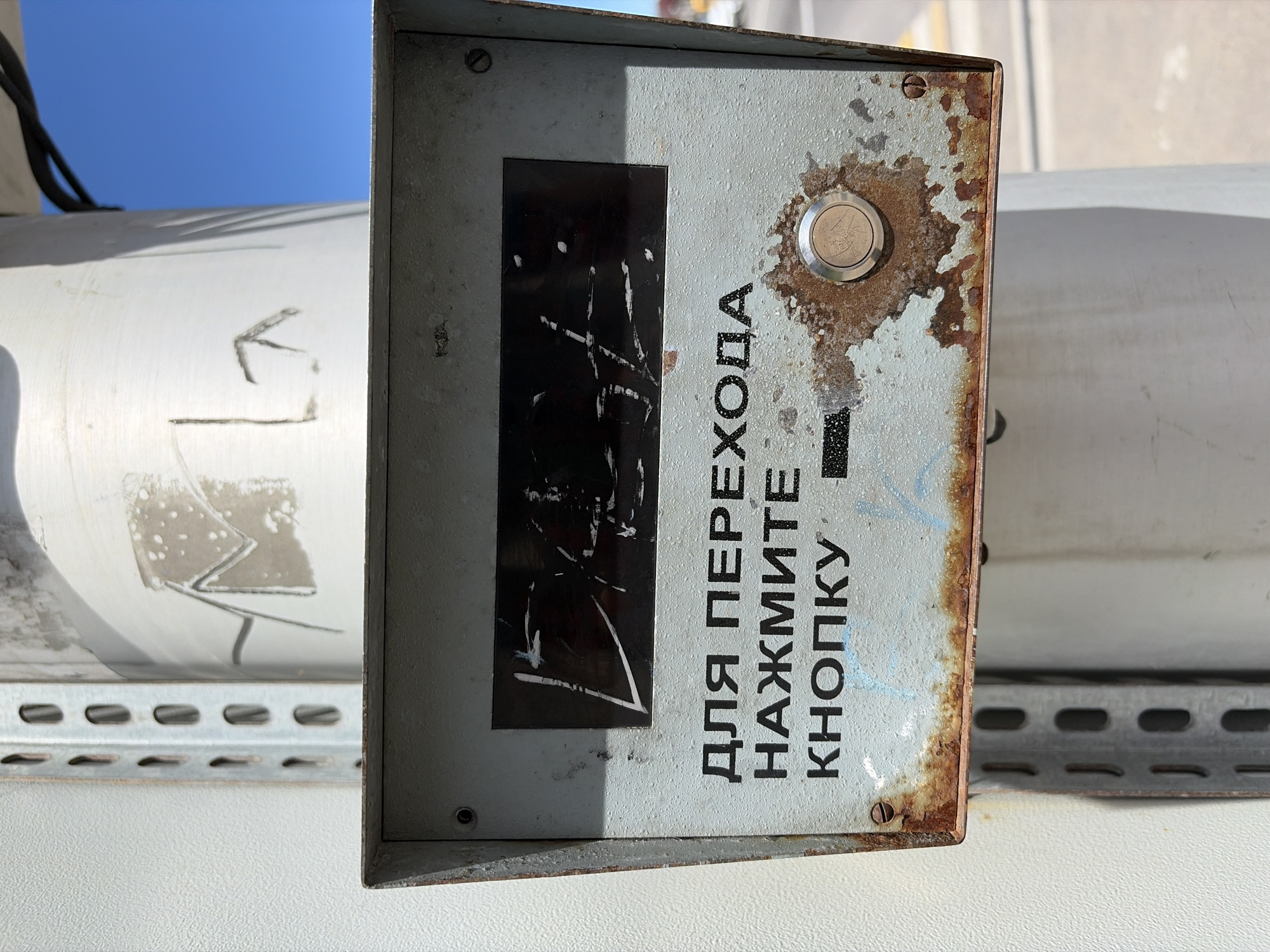 This is a close-up, eye-level shot of a weathered metal control panel, likely outdoors due to the bright sunlight casting a shadow on the left side of the panel. The panel is mounted on a cylindrical object, possibly part of a larger industrial or public utility apparatus. The sky, a clear blue, is visible in the upper left corner, suggesting a sunny day.
The control panel itself is heavily textured with rust and paint chips, giving it a worn and aged appearance. Centered on the panel is a black rectangular area that appears to be a label or a screen, marred with white scribbles and marks. To the right of this black area is a circular metallic button, also showing signs of wear.
Prominently displayed on the panel are Cyrillic characters in black text. The text reads: "ДЛЯ ПЕРЕХОДА НАЖМИТЕ КНОПКУ". This translates to "FOR TRANSITION PRESS THE BUTTON". The letters are bold and somewhat faded, consistent with the overall condition of the panel. Some faint blue graffiti or markings are also present on the panel, adding to its distressed look.
To the left of the main control panel, a portion of another large, cylindrical object is visible. This object also appears to be made of metal and has some markings, including what looks like a stylized arrow pointing to the left. Below this, a metal bracket with regularly spaced oval holes is visible, suggesting it's part of a structural support or mounting system. The overall impression is one of an aging, functional piece of equipment in an outdoor or industrial setting.