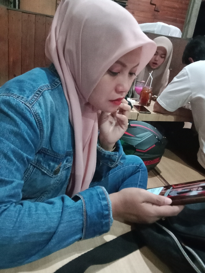 The image depicts an indoor scene, likely a cafe or restaurant, located in the city of Kebonsari, Indonesia. In the foreground, a woman wearing a light pink hijab, a blue denim jacket, and blue jeans is positioned facing downwards. Her left hand is near her mouth or chin, possibly applying or adjusting red lipstick, while her right hand holds a smartphone horizontally, displaying a screen. A black and red motorcycle helmet rests on the floor next to her. In the background, two other individuals are seated at a light-colored wooden table. One woman, wearing a light beige hijab, is partially visible, with two drinks—one pink, one brown—in glasses with straws on the table in front of her. Next to her, a man in a white short-sleeved t-shirt is visible from the back, also seated at the table. A second smartphone is visible on the table. The floor appears to be a light wood or laminate, with a dark strap or bag partially visible. The wall behind the subjects is constructed from vertical wooden planks.