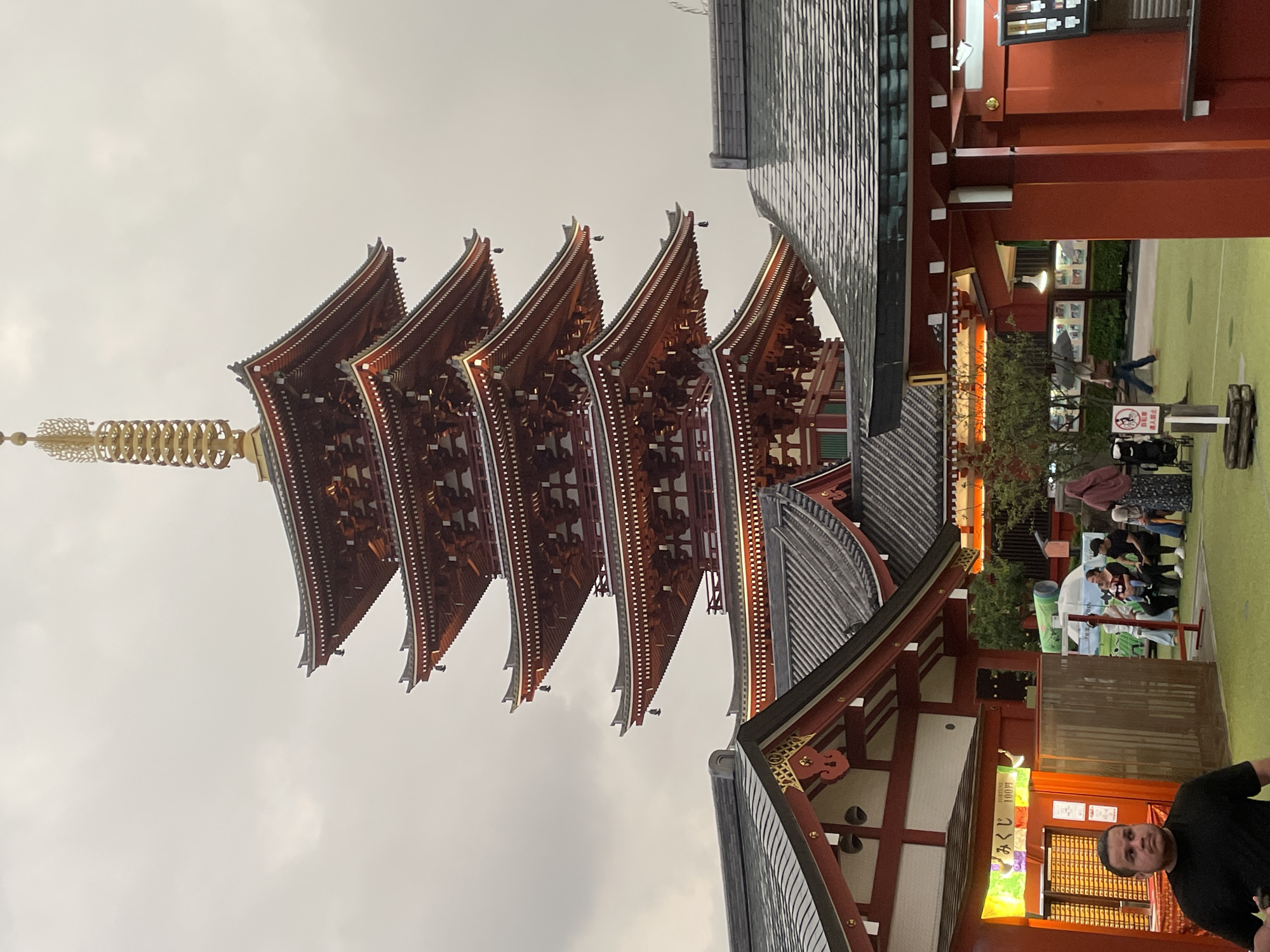 A five-tiered red pagoda stands tall against a hazy sky in Taito, Japan. The pagoda is adorned with gold accents and bells hang from its eaves. In the foreground, traditional Japanese buildings with dark tiled roofs and red trim surround a courtyard. Several people are visible, including a man in a black t-shirt walking towards the camera, and a group of people sitting together, one holding a child. A person with a headscarf is also present, as is another individual walking with an umbrella. A fortune-telling booth with a bright orange exterior is also visible. The overall scene suggests a cultural or tourist site within the city.