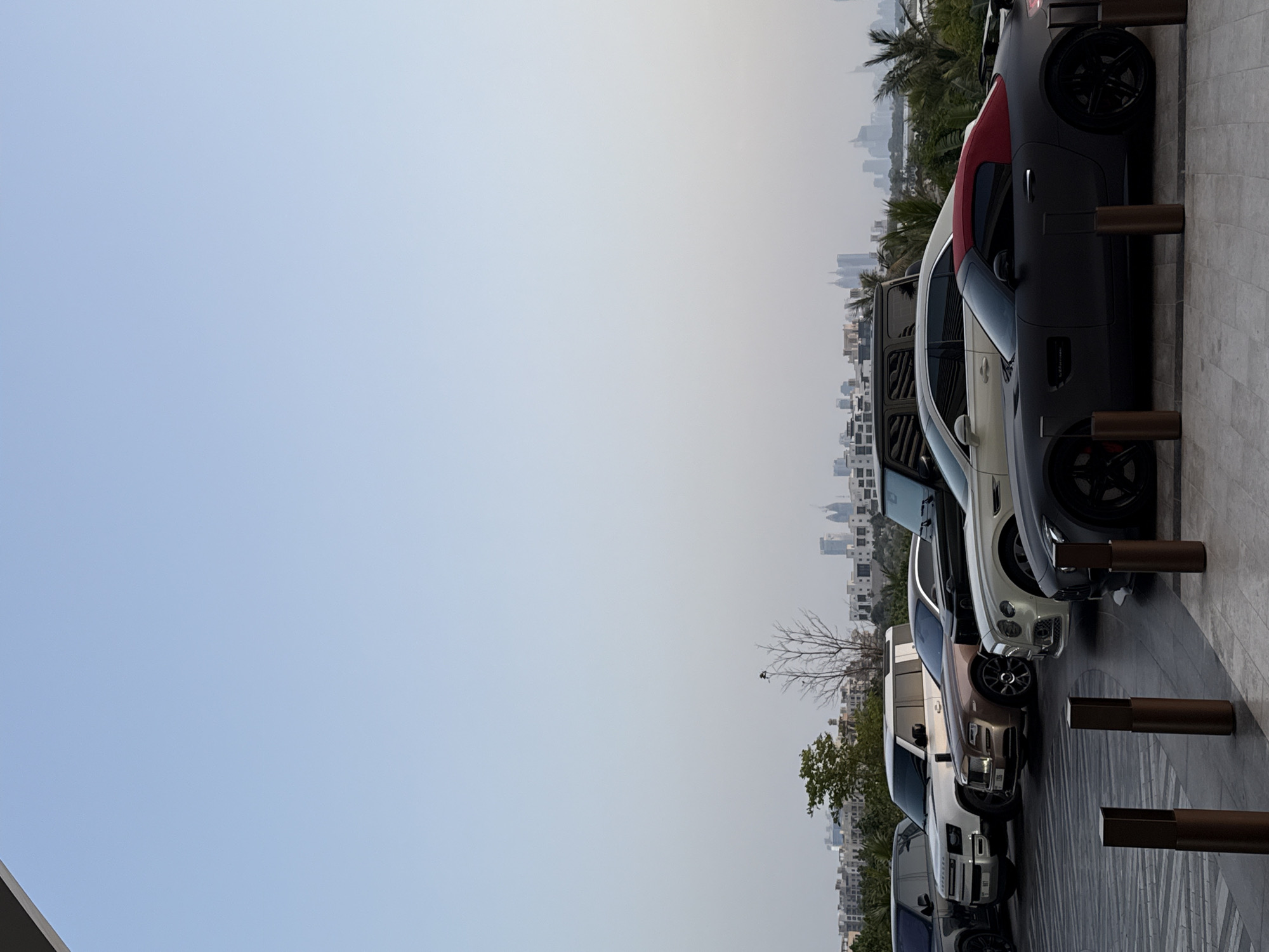 A row of luxury cars is parked on a dark, textured paved surface, extending from the mid-ground towards the foreground on the right side of the frame. The original image appears to be rotated 90 degrees counter-clockwise from a typical landscape orientation.
The vehicles, from left to right (as if viewed upright), include: a dark-colored SUV (likely a Land Rover Defender), followed by a light brown/bronze Rolls-Royce Cullinan SUV, then a cream/white Bentley (possibly a Continental GT or Flying Spur), and a dark grey matte-finish Mercedes-AMG GT Roadster with a distinctive red soft top and "V8 BITURBO" text visible on its front fender. Another dark-colored car is partially visible at the far right.
Several tall, rectangular, dark-colored bollards or decorative posts are evenly spaced along the edge of the paved area, to the right of the parked cars.
The background features a sprawling, hazy cityscape with numerous high-rise buildings and skyscrapers, indicative of a modern metropolis. Sparse greenery and palm trees are visible between the cars and the distant skyline. The left and upper portions of the image are dominated by a clear, pale blue sky, suggesting daytime conditions, possibly with atmospheric haze common in desert environments. The scene depicts a quiet moment with no human activity, highlighting the urban landscape and luxury vehicles common in Dubai, United Arab Emirates.