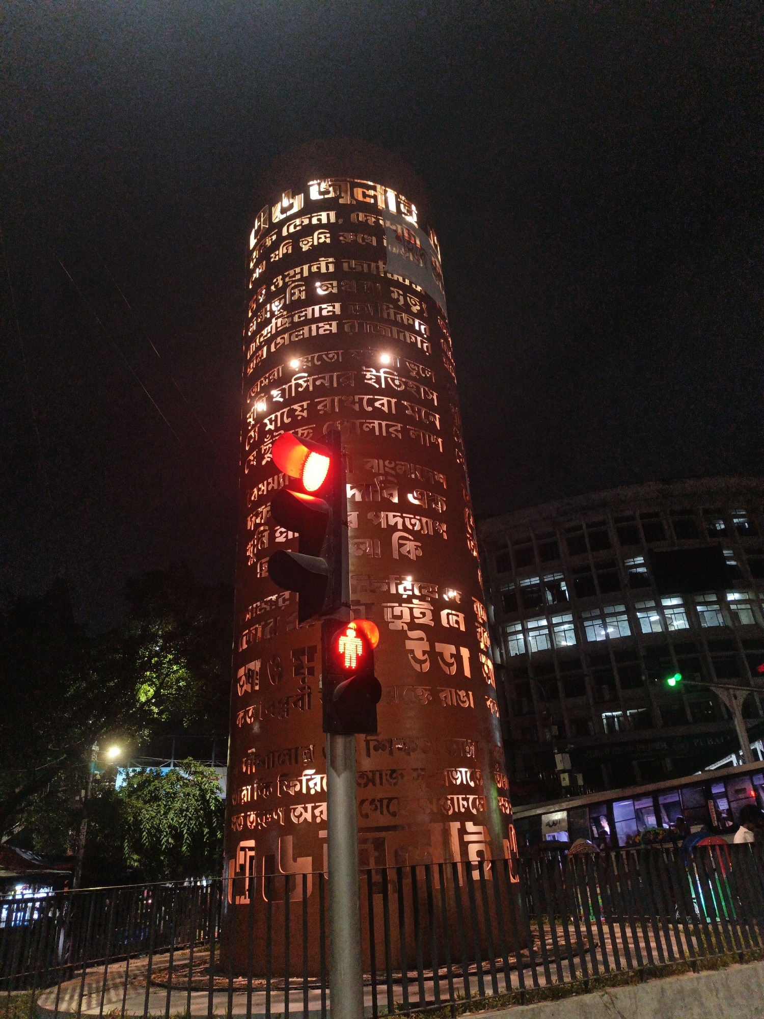 This is a nighttime photograph taken in Dhaka, Bangladesh. The main subject is a cylindrical monument, illuminated from within, with text cut out of its surface in Bengali script. The monument is located at a street intersection, evidenced by a traffic light. The traffic light has a red signal illuminated for vehicles and a red pedestrian signal showing a walking figure. In the background, a multi-story building with many lit windows is visible, suggesting an urban environment. Trees can be seen to the left of the monument. The scene is dimly lit, typical of a city at night, with the monument and building lights providing the primary illumination. There are no discernible people engaged in specific actions. The overall impression is that of a cityscape with a prominent cultural monument.
