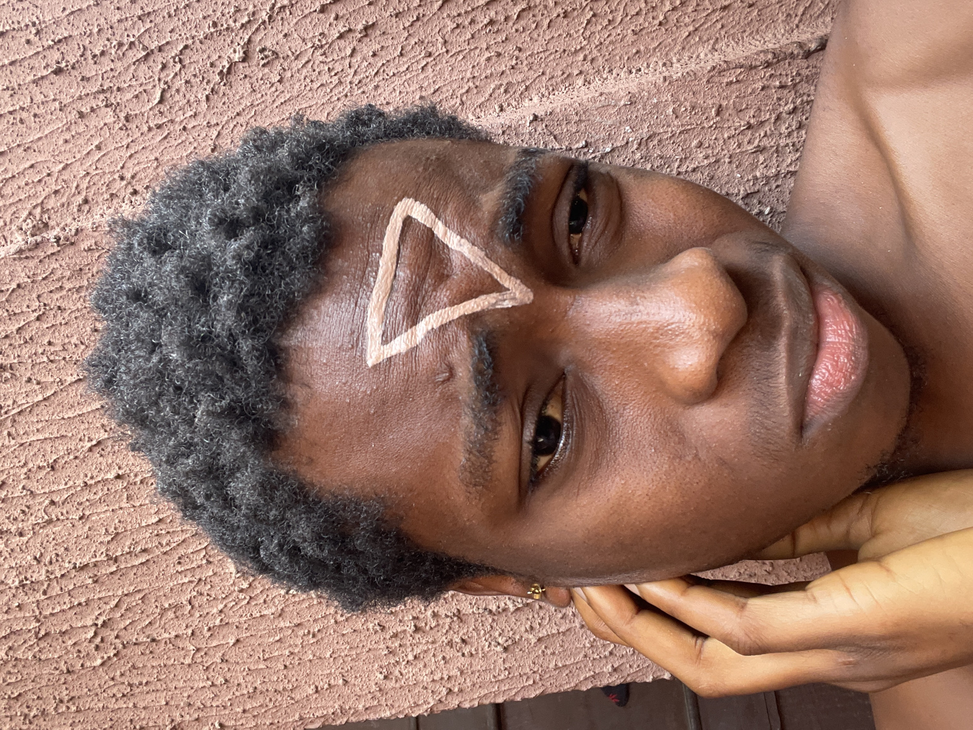 A medium close-up, slightly angled shot shows an individual with dark skin lying down, their head tilted slightly. A prominent white triangle is painted on their forehead, between their eyebrows. The individual has short, dark, tightly curled hair. The background appears to be a textured, tan-colored wall. The location is Nkpor, Nigeria. The individual's right hand is visible, cradling their face near the chin.
