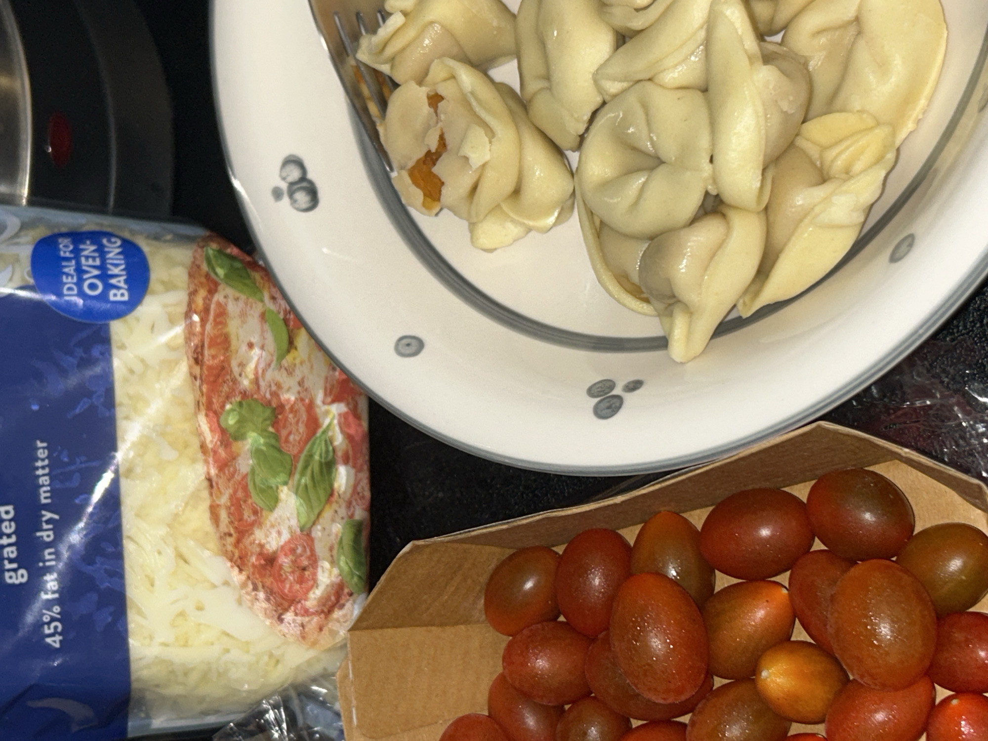 The image shows a close-up of food items, with no indication of a specific city or country. In the foreground, a cardboard container is filled with small, dark red cherry tomatoes. To the right, a white bowl with a gray rim and decorative gray circles contains several pieces of tortellini pasta. One piece of tortellini is partially open, revealing an orange filling. In the background, a clear plastic bag contains shredded cheese, with text on the bag indicating "grated 45% fat in dry matter" and "IDEAL FOR OVEN-BAKING."