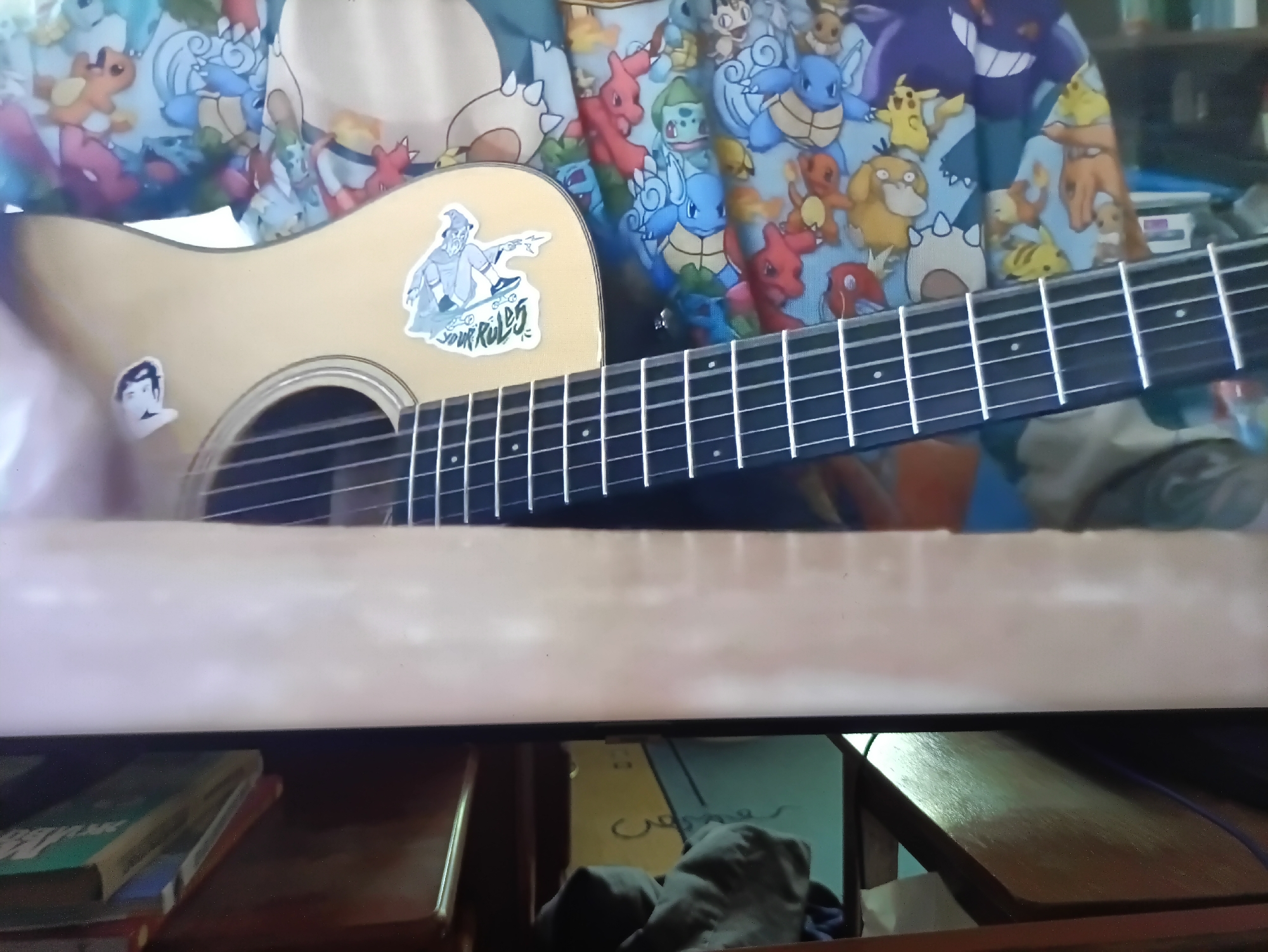 This is a close-up, eye-level shot of a person's hands and an acoustic guitar in what appears to be a home or studio setting. The guitar has a light-colored wood body and is adorned with stickers. The background is a colorful fabric with a repeating pattern of animated cartoon characters, specifically resembling characters from the Pokémon franchise. The fabric appears to be draped or worn as clothing. The guitar has metal strings and frets, and the tuning pegs are visible at the top. In the lower portion of the image, a horizontal band of light pink material runs across the frame, obscuring the lower part of the guitar and the immediate surroundings. Below this band, shelves are visible, holding books and other indistinct objects. The overall impression is of a casual, personal space where music is being played or practiced. The image does not contain any identifiable landmarks or elements that would allow for its location to be determined within the city of Kamianets-Podilskyi, Ukraine.