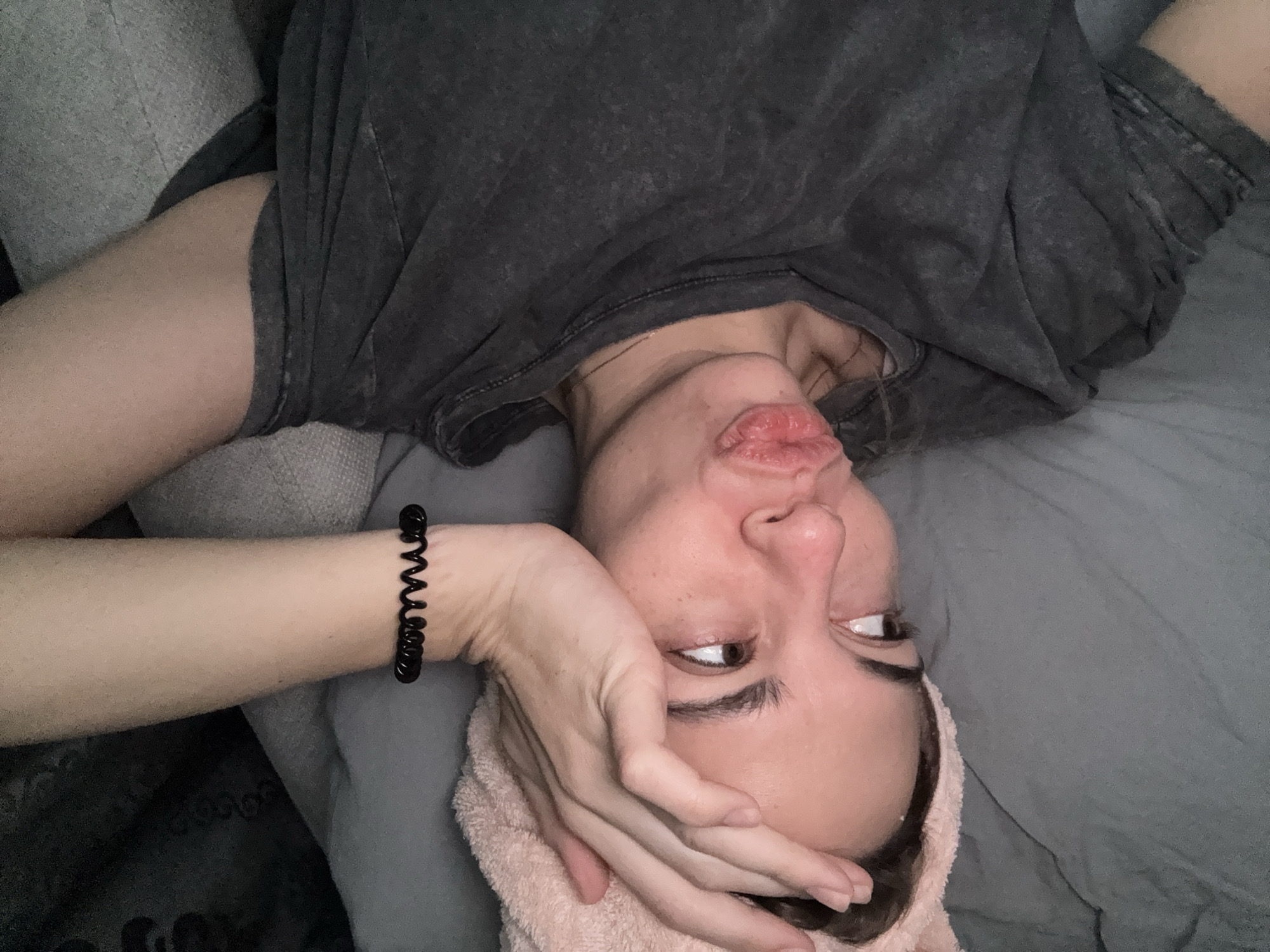 A woman lies supine on a gray bed with gray bedding. Her head is positioned at the bottom of the frame. She wears a gray t-shirt, and a pink towel is wrapped around her head. Her left arm is raised, her hand partially obscuring her face. A black spiral hair tie is visible on her left wrist. The woman's lips are pursed. The background is a mix of gray fabric and indistinct shapes. There is no information in the image to indicate that this scene is in Ust-Ilimsk, Russia.