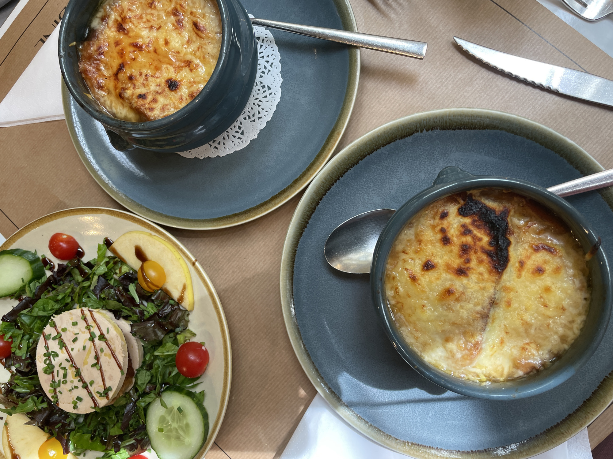This is a top-down, brightly lit shot of a table set for a meal in what appears to be a French restaurant, aligning with the provided context of Paris, France.
The scene features three dishes arranged on a light brown, possibly paper, tablecloth. Two of the dishes are classic French onion soups, served in dark grey ceramic bowls with handles. Each soup is topped generously with golden-brown, melted, and gratinéed cheese and croutons. One bowl sits on a white paper doily atop a matching grey plate, with a silver spoon to its left. The other soup bowl, also on a matching grey plate, has a spoon resting inside it.
The third dish is a vibrant salad, presented on a light-colored plate with a brown rim. The base of the salad consists of mixed green leaves, adorned with red and yellow cherry tomatoes, sliced cucumbers, and thin slices of apple. Central to the salad are two circular servings of what appears to be pâté or foie gras, garnished with chopped chives and drizzled with a dark, glossy sauce, likely a balsamic reduction.
Silver cutlery, including a serrated knife and another spoon, is visible on the tablecloth. A white paper napkin is partially visible beneath one of the plates. In the upper left background, part of a word "DINE" is visible on a piece of paper or menu, further indicating a restaurant setting. The overall presentation suggests a casual yet inviting dining experience, showcasing typical French cuisine.