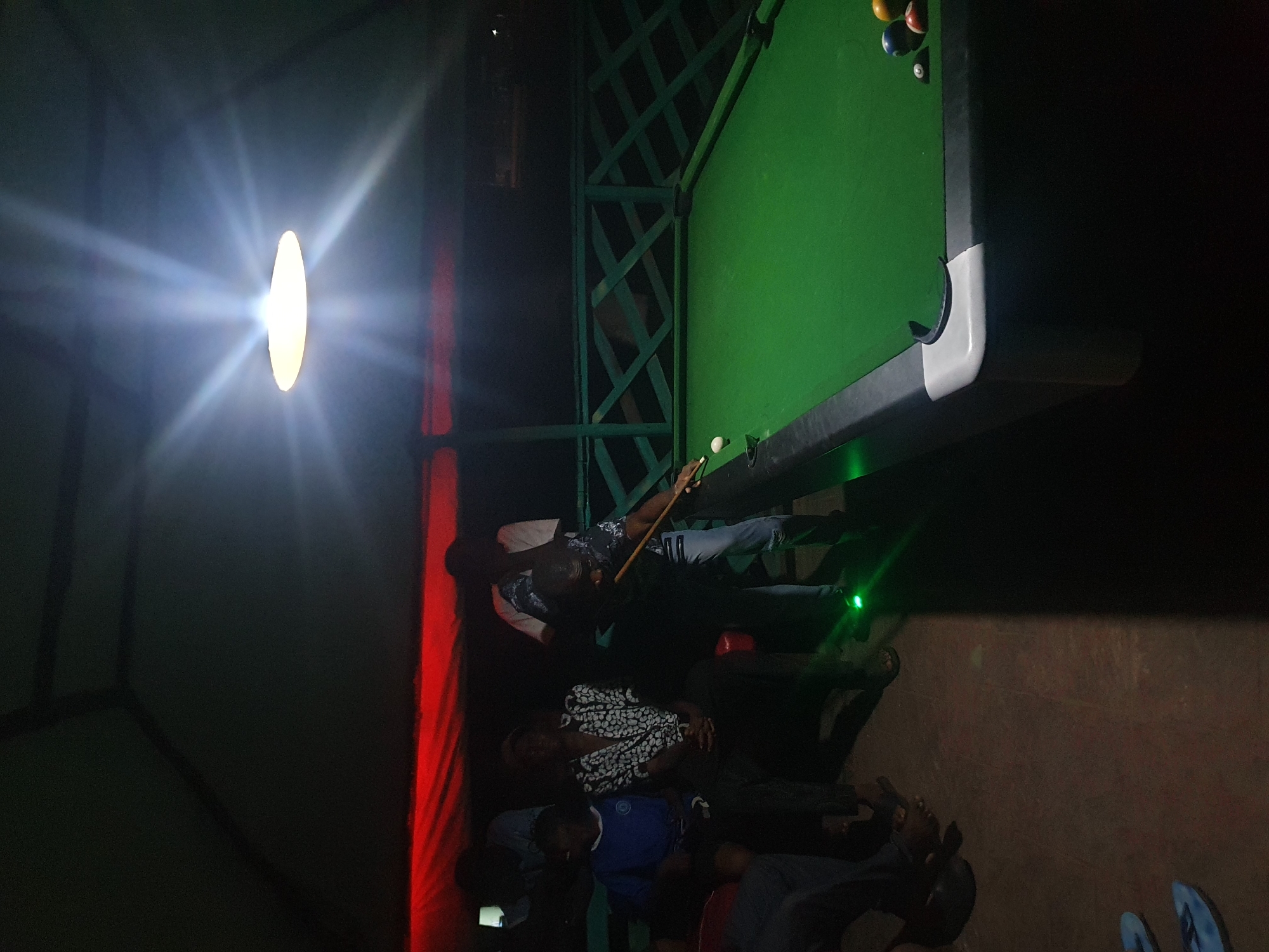 A group of men are gathered around a pool table at night, possibly in an outdoor recreational area under a covered structure. The pool table is green with black and white accents. Several pool balls are visible on the table. One man, wearing a patterned shirt and jeans, is actively playing pool, holding a cue stick and appearing to strike a ball. Other men are standing or sitting nearby, observing the game. One man in a blue shirt is seated. The environment is dimly lit by overhead lights, with a bright light source directly above the pool table. There is a red-colored element visible in the background, potentially lighting or a decorative element. A green railing or fence is also visible behind the pool table. The scene suggests a social gathering or a recreational activity taking place in Lokoja, Nigeria.