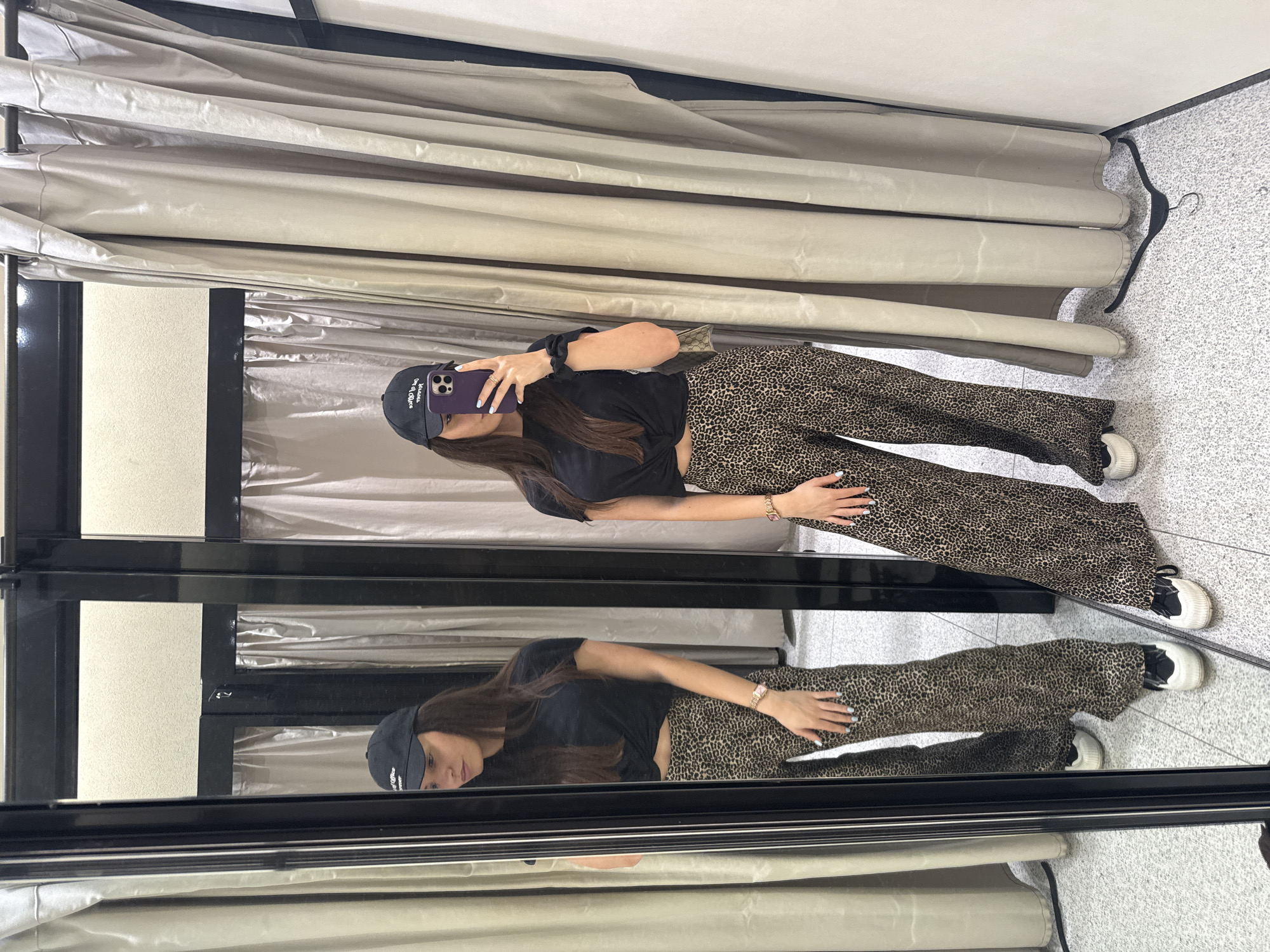 A woman stands in a dressing room, taking a mirror selfie with a purple iPhone. She is wearing a black baseball cap, a short-sleeved black shirt, leopard print pants, and white sneakers with black accents. She has a bracelet on her left wrist. The dressing room features a black-framed mirror, beige curtains, and a gray speckled floor. A black hanger is visible on the right side. The location is a retail store in Plovdiv, Bulgaria.
