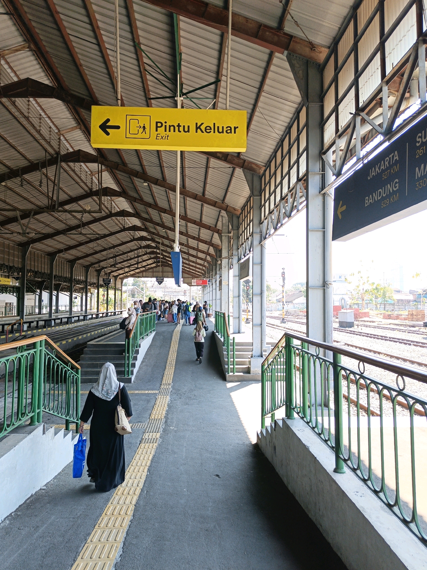 This is a daytime, eye-level, outdoor, medium shot of a covered train station platform in Surakarta, Indonesia. The platform is paved with concrete and features a yellow tactile paving strip running along the edge. Green metal railings border the platform. A sign with a yellow background and black text indicates "Pintu Keluar" (Exit) with an arrow pointing left. Another sign, dark gray with white text, lists destinations and distances to Jakarta (527 km) and Bandung (529 km).
Several people are visible on the platform. In the foreground, a person wearing a dark abaya and headscarf walks away from the camera, carrying two bags, one blue and one beige. Further down the platform, a group of people are standing, some appearing to be waiting for a train. One individual in the group is wearing a light-colored headscarf and looking towards the tracks.
The station has a metal roof supported by columns, with a corrugated metal roof. Natural light streams in from the open sides, casting shadows. Train tracks are visible beyond the platform. The overall impression is of a functional, active transportation hub under bright daylight.