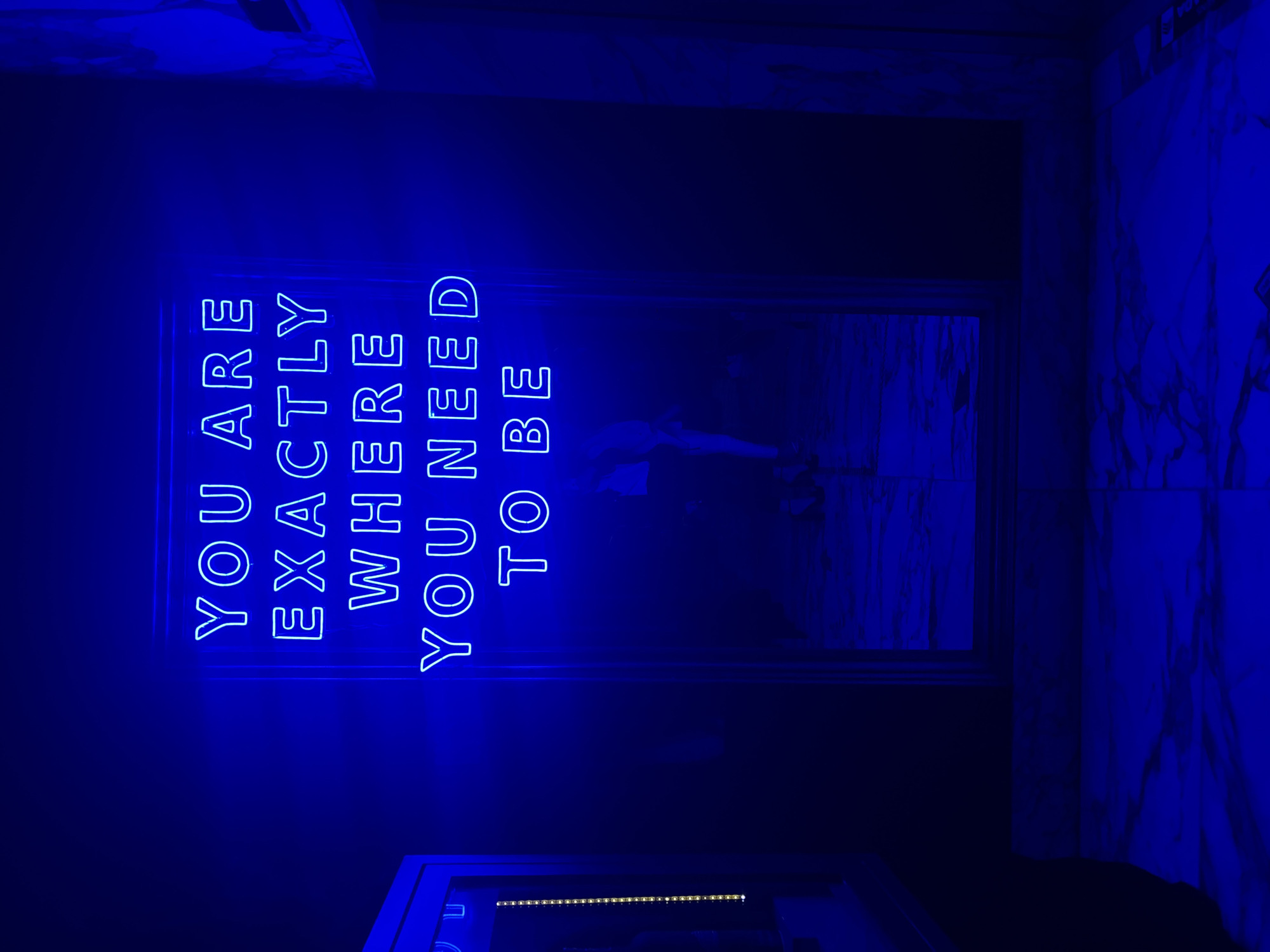 The scene depicts an interior space in Mexico City, Mexico, dominated by a vertically oriented neon sign glowing bright blue against a dark background. The sign displays the capitalized text: "YOU ARE EXACTLY WHERE YOU NEED TO BE" across five lines. Below the neon sign, a dark, reflective surface, likely a mirror or polished floor, shows a blurry, indistinct silhouette of a person, possibly bending or standing. The reflective surface also captures the blue light from the neon, extending its glow onto a tiled floor in the foreground. These tiles are large, light-colored, and appear to have a subtle, swirling pattern, all rendered in shades of blue due to the ambient neon illumination. To the far left, a framed vertical element with a strip of small, yellowish-white lights is partially visible, providing a contrasting warm tone to the predominantly cool blue environment. The overall lighting is dim, with the neon sign serving as the primary light source, casting a deep blue hue across the visible surfaces.
