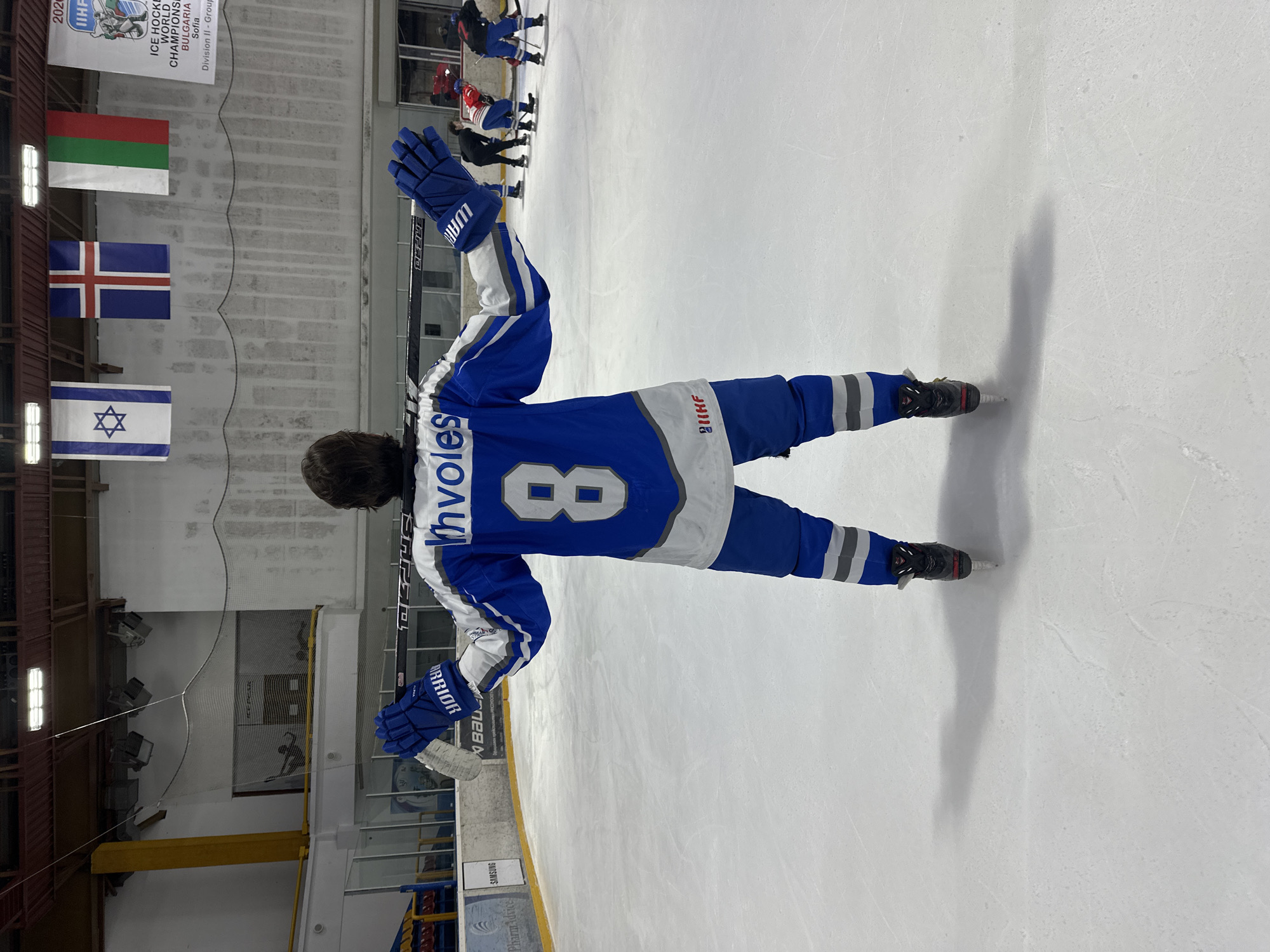 The image shows a person in a blue and white hockey jersey with the number 8 on the back and the name "Ivoves" above it. The person is standing on an ice rink, wearing hockey skates and gloves, and holding a hockey stick. Other players are visible in the background. Banners displaying the flags of Bulgaria, Iceland, and Israel hang on the wall. A banner for the "2020 IIHF Ice Hockey World Championships, Bulgaria, Sofia, Division II - Group" is also visible. A sign that says "SAMSUNG" and "Pharm Advice" is visible in the bottom left corner. The scene is an ice hockey rink in Sofia, Bulgaria, during an IIHF World Championship event.