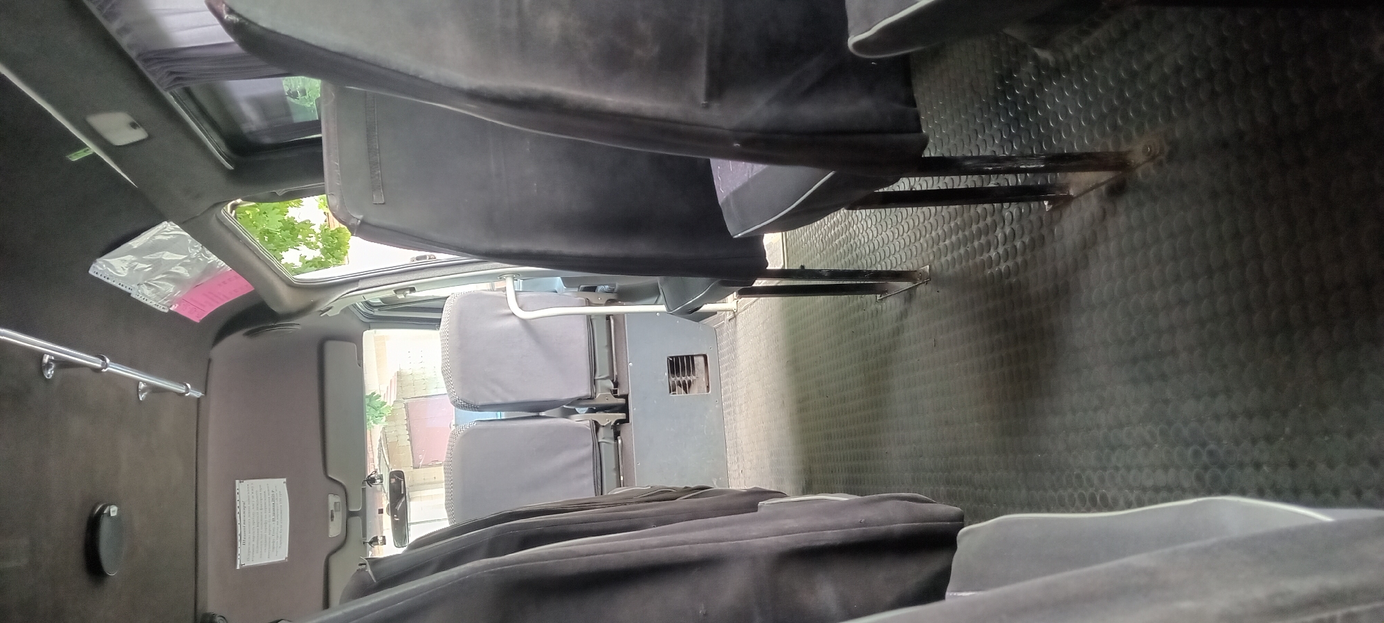 The image shows the interior of a passenger bus, likely parked outdoors on a sunny day, as indicated by the bright natural light filtering in from the windows and the visible trees and buildings outside. The bus has a dark grey, textured floor. There are rows of dark grey fabric-covered seats with metal frames. The seats in the foreground are partially obscured by the dark fabric covering of the seats in the front passenger area. In the middle ground, two grey fabric-covered seats are visible, facing forward. Between these seats, a grey panel with a black ventilation grate is mounted at the base. The ceiling of the bus is dark brown and appears to be upholstered. A metal bar runs along the ceiling. A white sign with black text is attached to the ceiling on the left side. On the right side, above the window, a clear plastic bag and a pink item are attached to the ceiling. The overall condition of the bus interior appears to be used but functional. There are no people visible in the image. The location is specified as Koviahy, Ukraine.