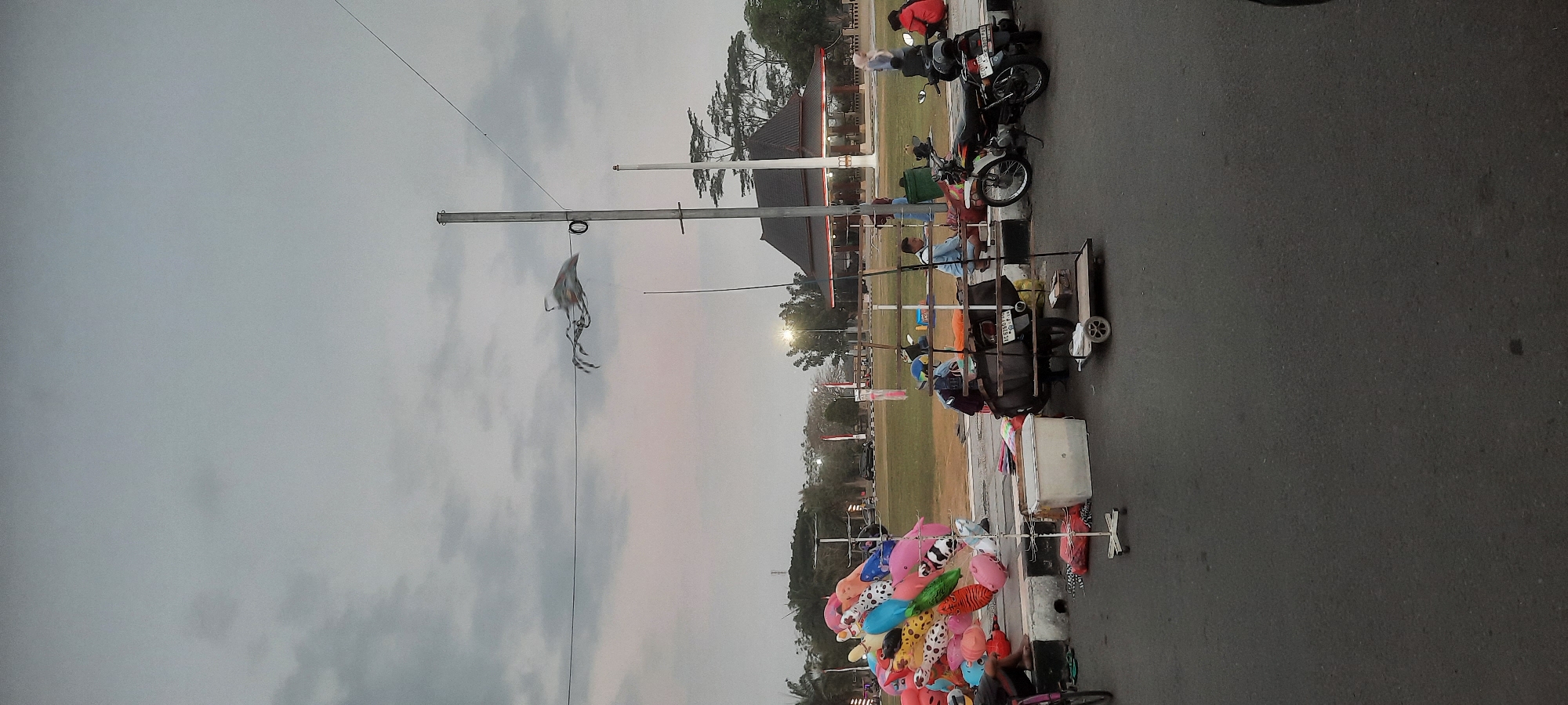 The image captures a street scene in Kanigoro, Indonesia, likely during the late afternoon or early evening, indicated by the overcast sky and artificial lighting in the distance. In the foreground, a sidewalk is lined with a collection of colorful balloons, suggesting a vendor is present or has recently been. Next to the balloons, a white cooler and a display rack with merchandise are visible. Several people are gathered near the sidewalk, some seated and others standing, with at least two motorcycles parked nearby.
Further back, a grassy field is visible, with trees and a building featuring a dark, tiered roof in the background. A tall utility pole with wires stretches across the sky, and a kite is suspended from one of the wires, appearing to be a traditional Indonesian kite, possibly a "layang-layang". The overall atmosphere is that of a casual gathering or a market setting on the edge of an open area. The road in the immediate foreground is asphalt, and the lighting conditions suggest low light.