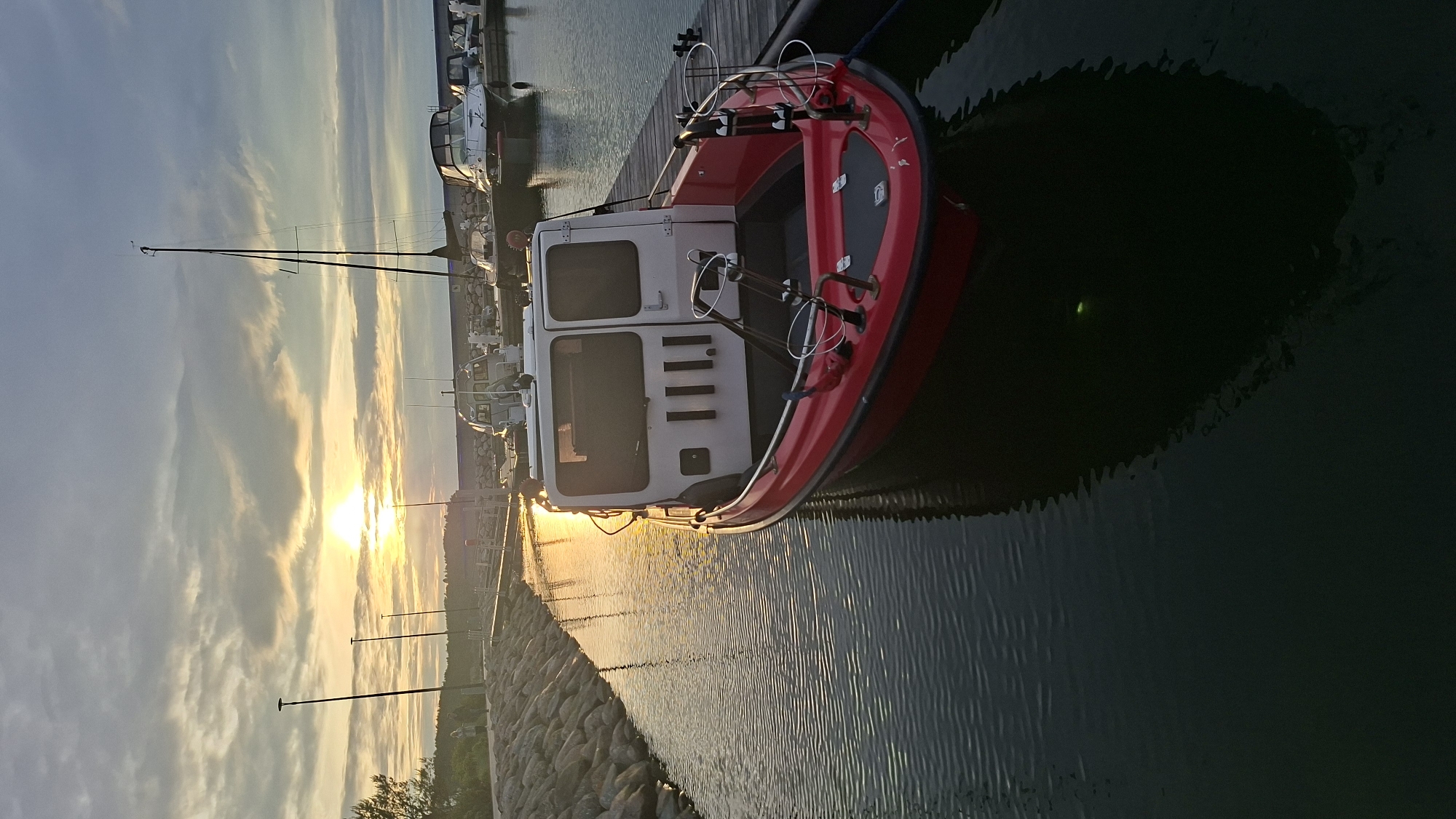 A red and white boat is docked at a harbor during sunset. The sun is low on the horizon, casting a warm golden light across the sky and water, with some clouds visible. The boat has a white cabin with windows and a red hull. It is moored to a wooden dock. In the background, other boats and a mast are visible. The water is calm, with ripples reflecting the light. The scene appears peaceful and serene, suggesting a quiet evening in the harbor. No people are visible. There is no visible text in the image. The location context is Tallinn, Estonia.