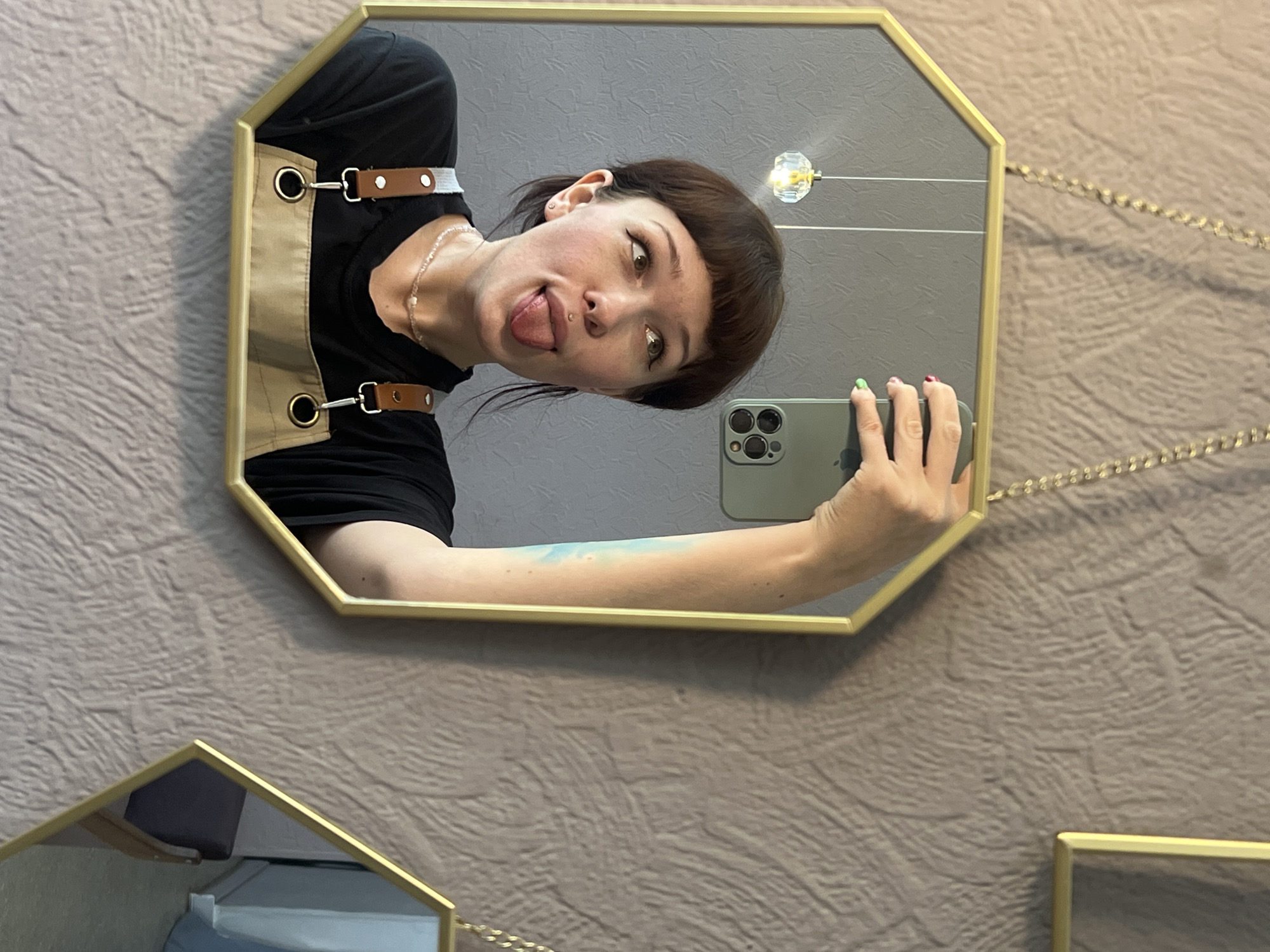 An octagonal, gold-framed mirror, suspended by a gold chain, captures a mirror selfie in an interior setting in Chaikovsky, Russia. A person with short, dark brown hair and blunt bangs is visible from the chest up, making a playful expression with their tongue extended and eyes looking upward. They have a philtrum piercing and an earlobe piercing. The individual is wearing a black t-shirt and a beige apron with brown leather straps and metallic hardware. A beaded necklace, predominantly pink and white, is visible around their neck. Their right hand, with fingernails painted in red and green, holds a dark green smartphone, which has a distinct multi-lens camera array, positioned to capture the reflection. A subtle blue marking or tattoo is visible on the person's right upper arm. The reflection in the mirror shows a solid gray wall with a small, crystal-like light fixture hanging from the ceiling. Outside the mirror's frame, the background consists of a textured, light brown wall. A partial view of another gold-framed hexagonal mirror is visible on the right side of the image, reflecting a dark object.