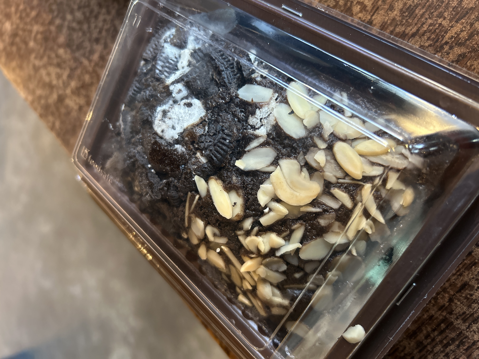 A close-up, high-angle shot shows a rectangular brownie in a clear plastic container, placed on a wooden surface. The brownie is dark brown and topped with sliced almonds and what appear to be crushed cookies or dark chocolate chunks. Some white powdery substance is visible on top of the brownie. The container has a brown base and a clear lid. The background is blurred, showing hints of what could be a surface with a light, muted texture. The image was taken in Sukawati, Indonesia.