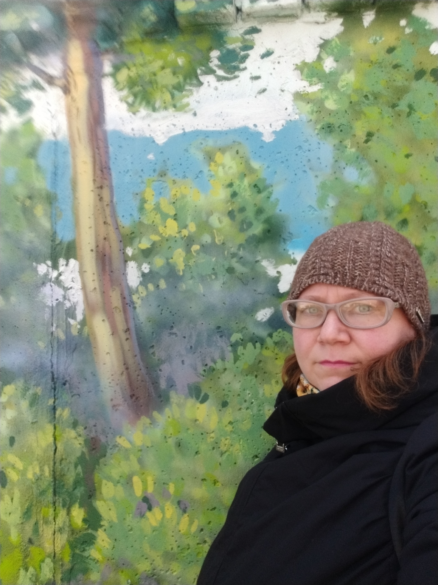 A medium shot depicts a woman wearing glasses and a brown knit hat, with a black coat pulled up around her neck. Behind her is a mural painted on a wall. The mural depicts a tree trunk with green and yellow foliage above, and a cloudy blue sky visible through the leaves. There are visible raindrops or water stains on the mural's surface. The photograph was taken outdoors in the city of Chaikovsky, Russia.